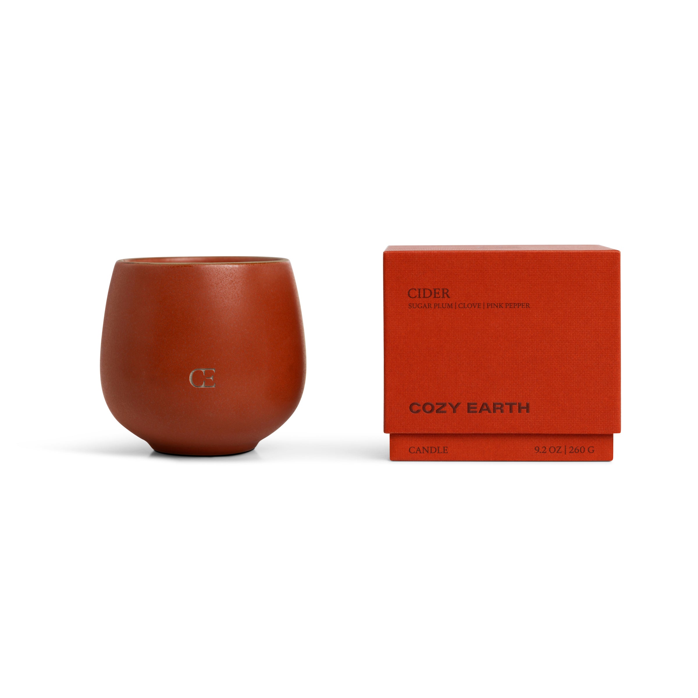 A brown, rounded Candle CIDER holder by Cozy Earth with “CE” embossed sits beside a matching red box labeled "Cider" and "Cozy Earth.” The box lists notes of sugar plum, clove, and pink pepper.