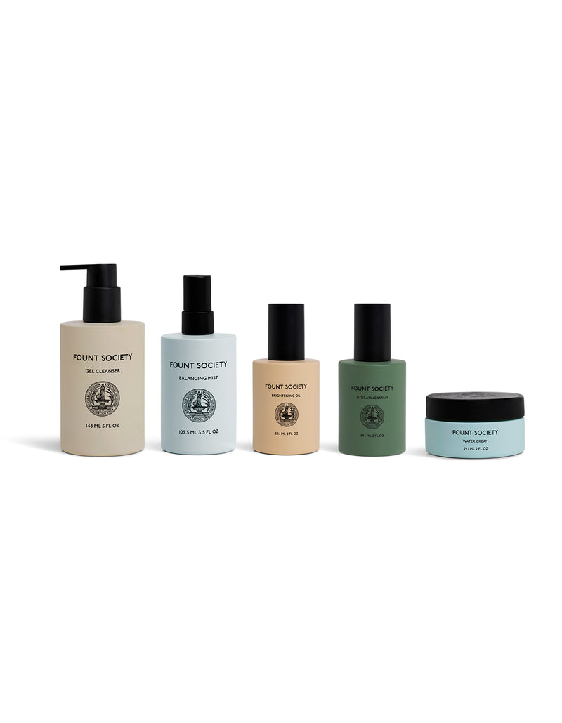 Five skincare products from Cozy Earth’s The Routine are arranged in a row, including a gel cleanser with a pump, a balancing mist spray, two bottles with black caps, and a small blue jar with a black lid.