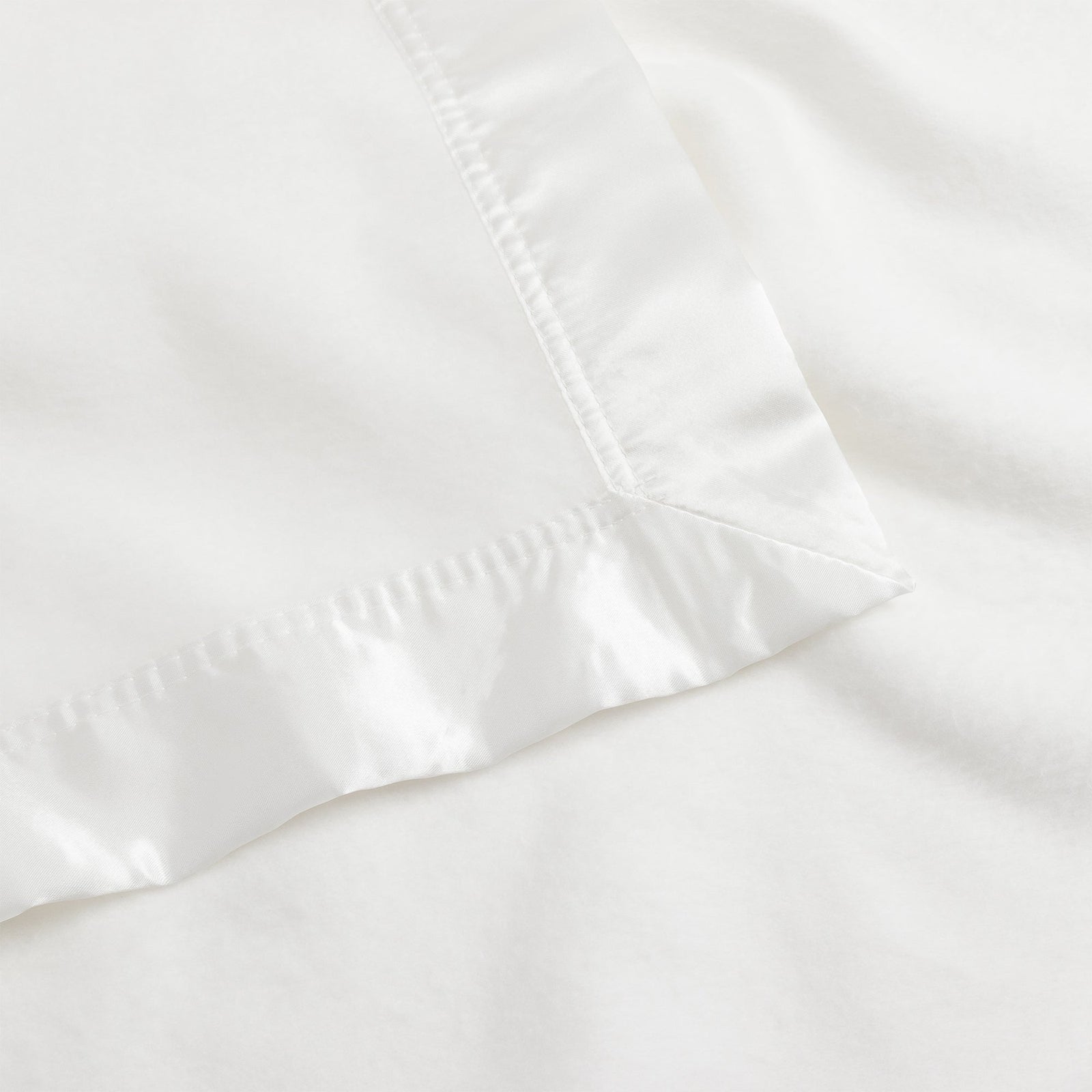 Close-up image of the Bamboo Blanket by Cozy Earth, featuring a white fabric with a satin border. The image highlights a neat corner with a smooth, glossy texture on the border and a matte, soft texture on the main fabric. 