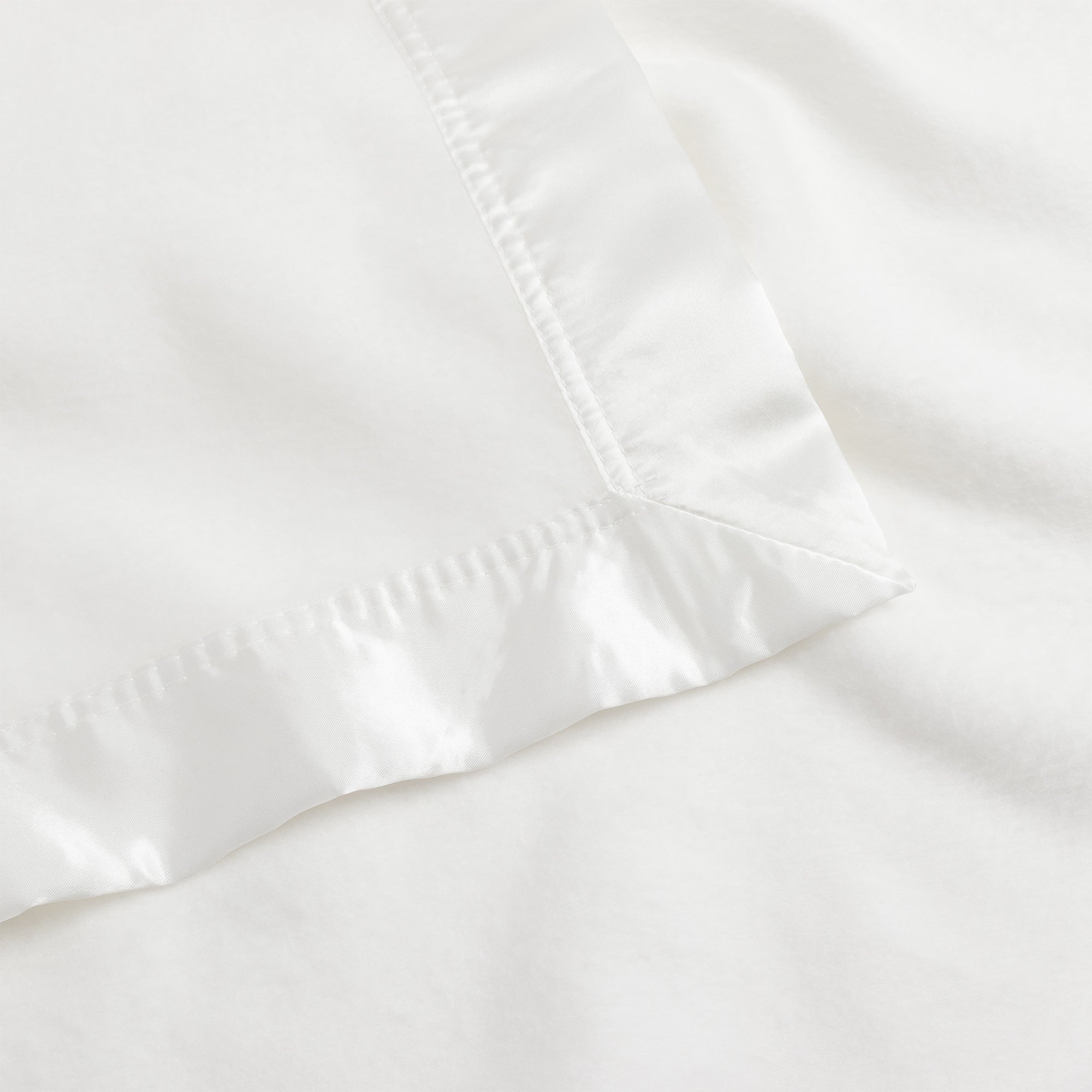 Close-up image of the Bamboo Blanket by Cozy Earth, featuring a white fabric with a satin border. The image highlights a neat corner with a smooth, glossy texture on the border and a matte, soft texture on the main fabric. |Color:White