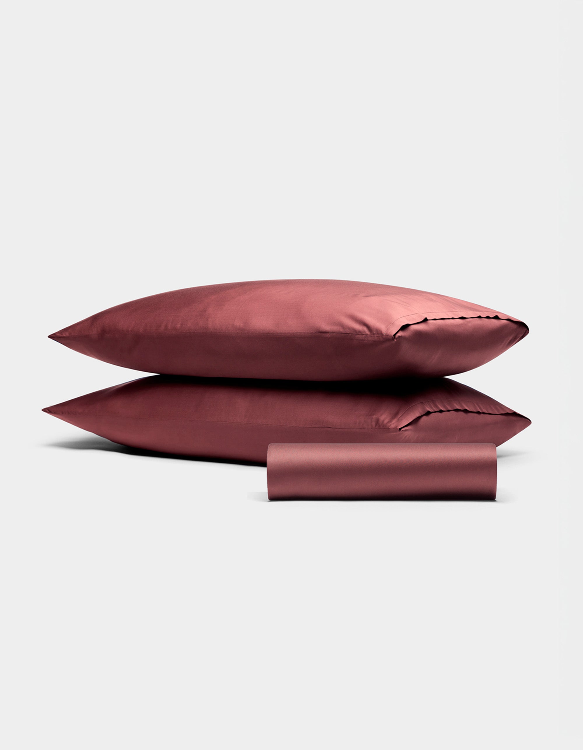 Two stacked, burgundy Bamboo Bedding Basic Bundle pillowcases by Cozy Earth with a matching rolled-up sheet, all set against a plain light gray background.|Color:Mulberry