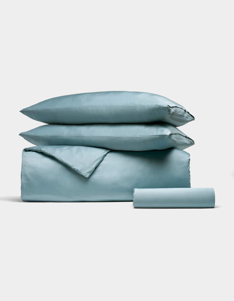 The Cozy Earth Bamboo Bedding Essential Bundle, featuring a neatly folded light blue set with two pillows, a duvet, and a rolled sheet, is stacked together on a simple light background.
