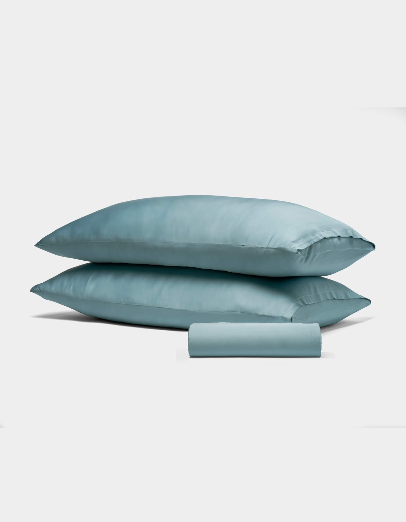 The Bamboo Bedding Basic Bundle by Cozy Earth features two light blue satin pillows stacked together, with a matching satin pillowcase rolled up in front, all displayed against a plain white background.