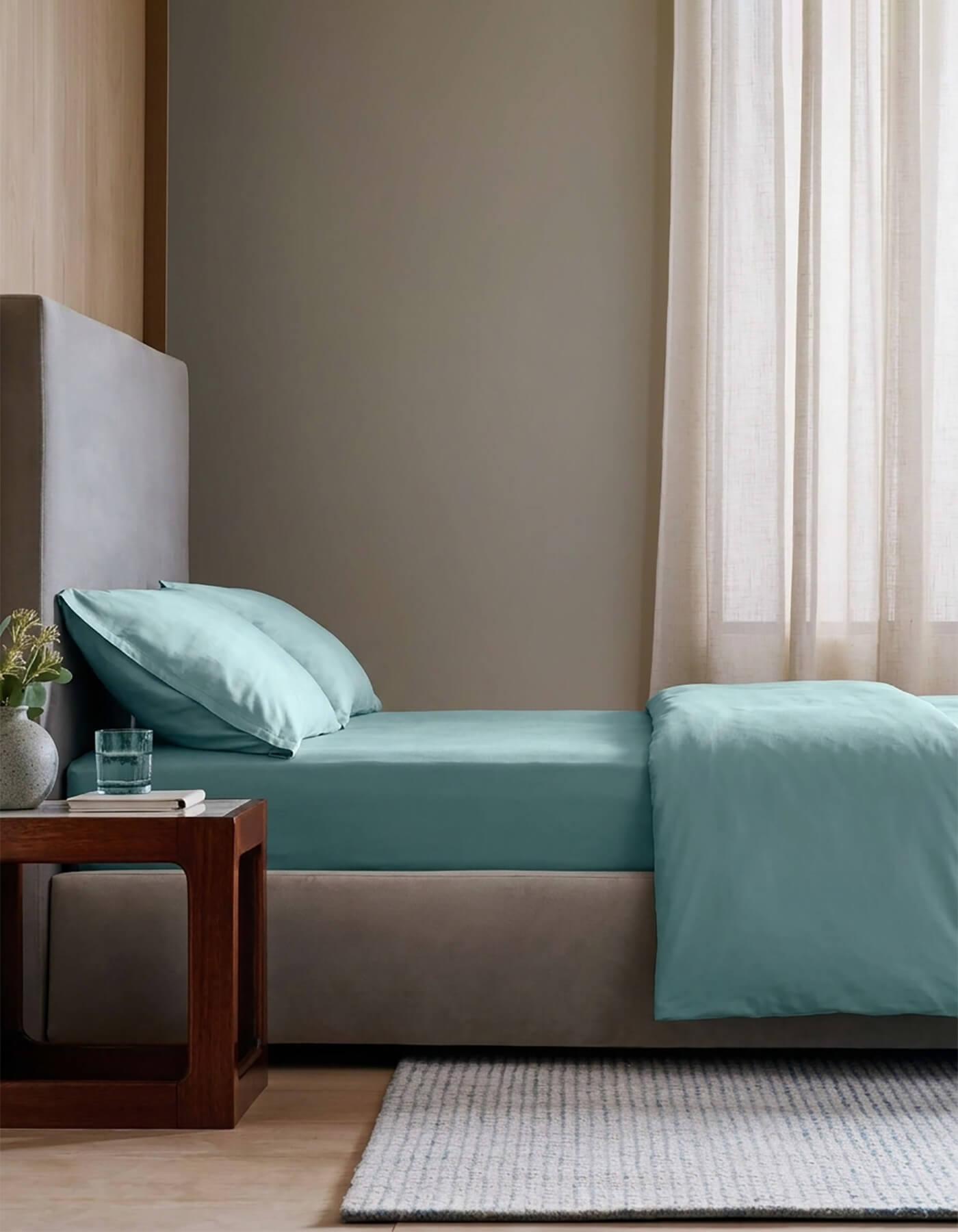 A modern bedroom with a neatly made bed featuring the Cozy Earth Bamboo Bedding Essential Bundle, light blue sheets and pillows, a wooden nightstand with a glass of water and plant, beige walls, and sheer curtains letting in natural light.