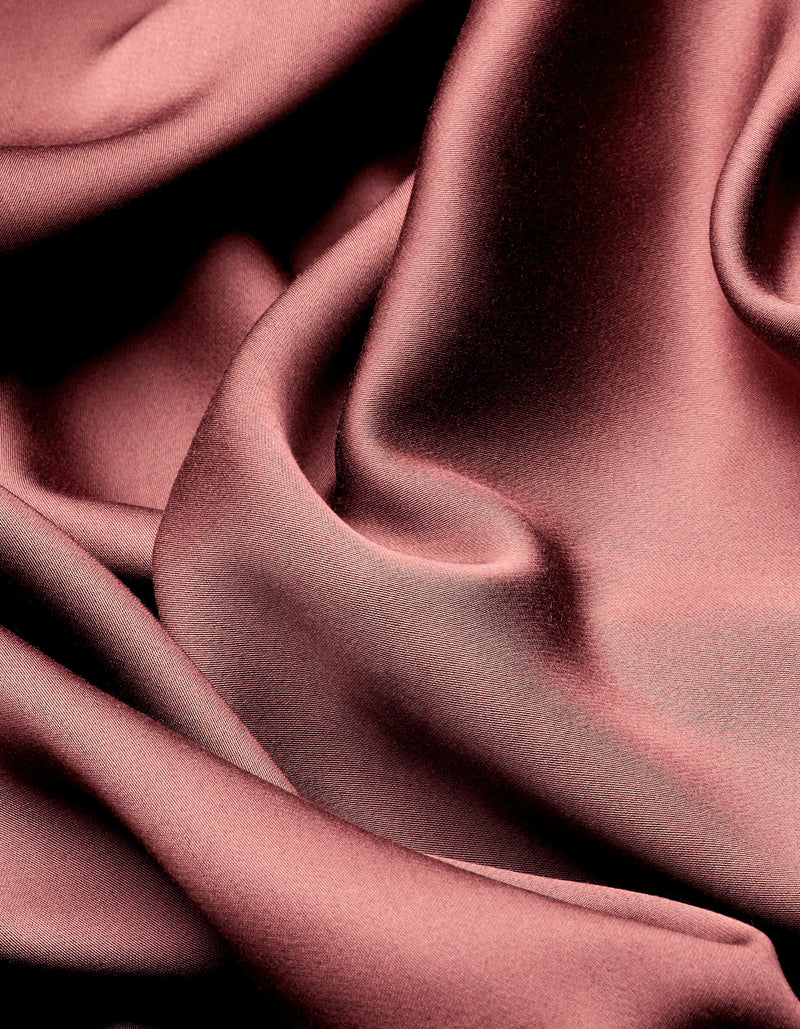 Smooth, shiny Bamboo Sheet Set by Cozy Earth drapes in soft, flowing folds, creating elegant highlights and shadows across the rose-colored satin surface.