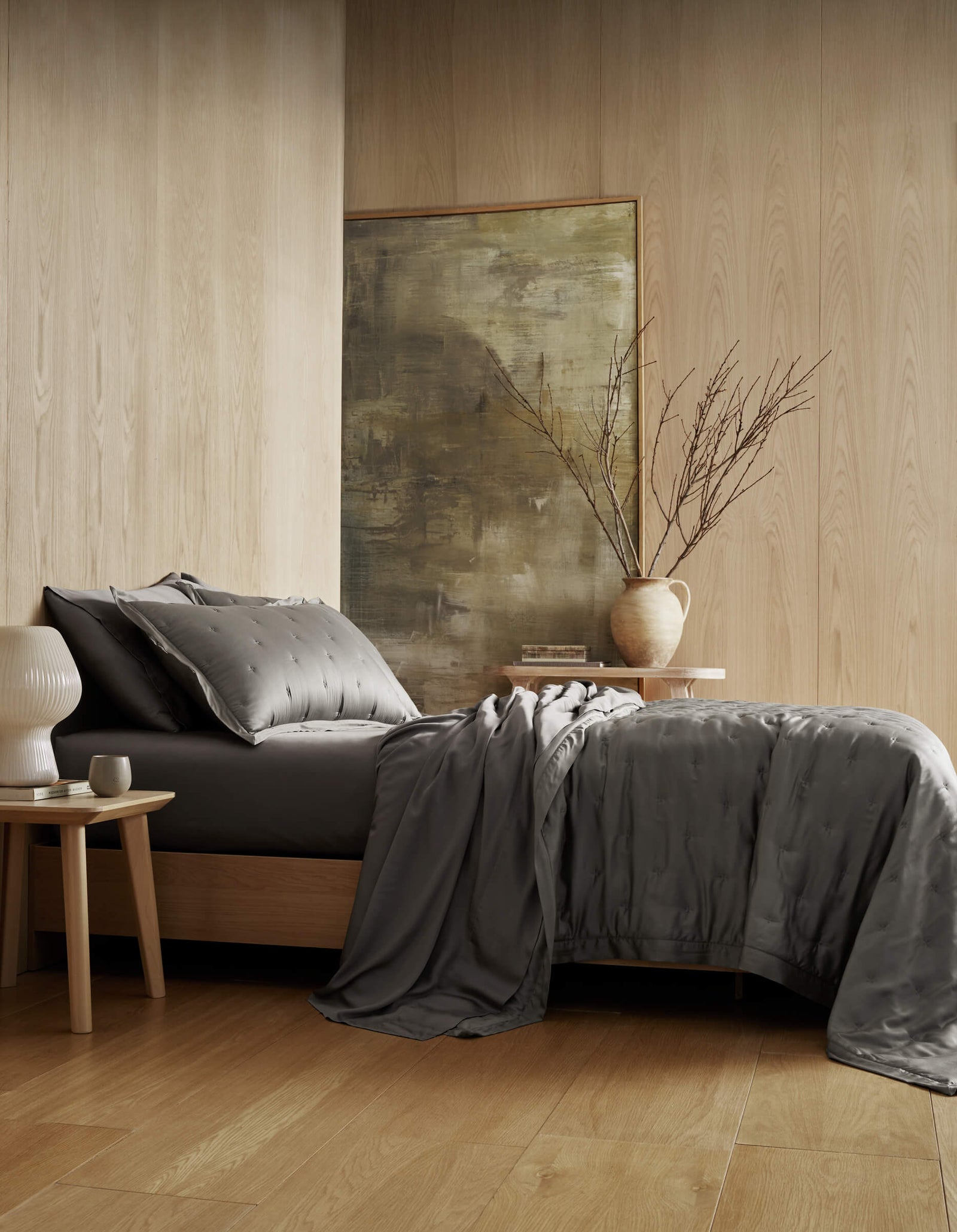 A modern bedroom with light wood walls and floors features a bed dressed in the Cozy Earth Bamboo Quilt in dark gray, a nightstand with a white lamp, another table with a vase of branches, and abstract artwork in the background.