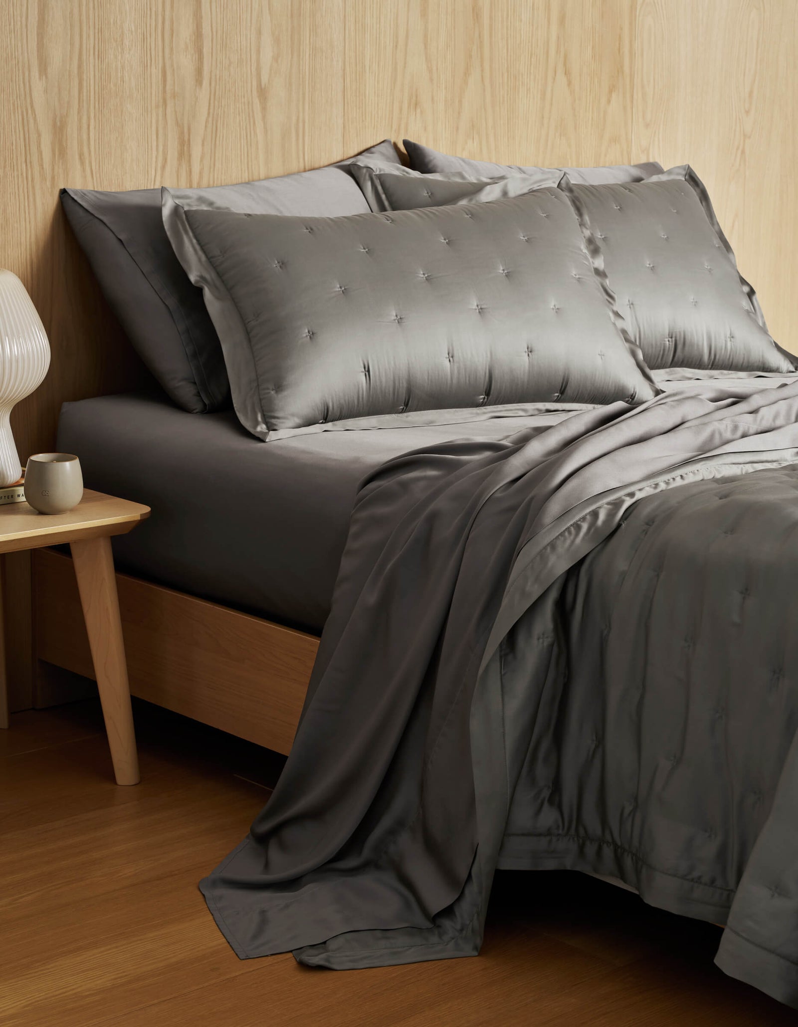 A modern bedroom features a Cozy Earth Bamboo Quilt with gray silk bedding and matching pillows on a wooden bed frame, beside a wooden nightstand with a white ceramic lamp and cup, set against a light wood-paneled wall.