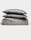 The Bamboo Quilt Bundle by Cozy Earth features lustrous silver-gray bed linens, including two stacked pillows, a folded sheet, and a duvet cover, all arranged against a plain white background.|Color:Charcoal