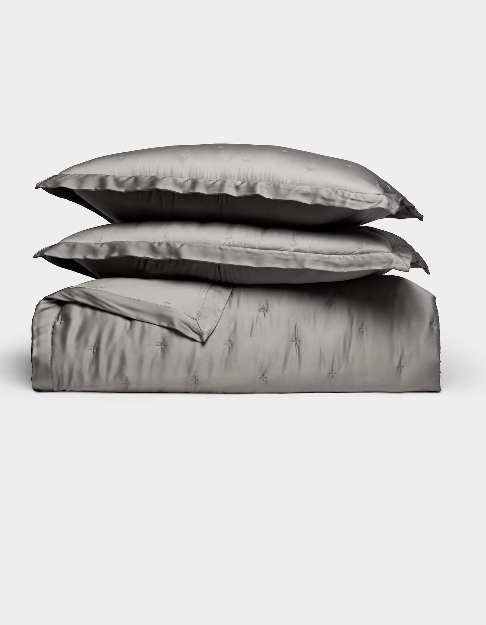 The Bamboo Quilt Bundle by Cozy Earth features lustrous silver-gray bed linens, including two stacked pillows, a folded sheet, and a duvet cover, all arranged against a plain white background.