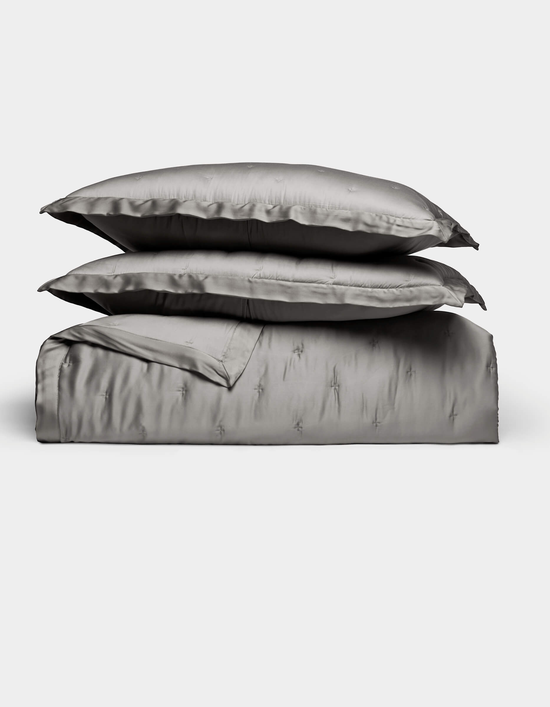 The Bamboo Quilt Bundle by Cozy Earth features lustrous silver-gray bed linens, including two stacked pillows, a folded sheet, and a duvet cover, all arranged against a plain white background.