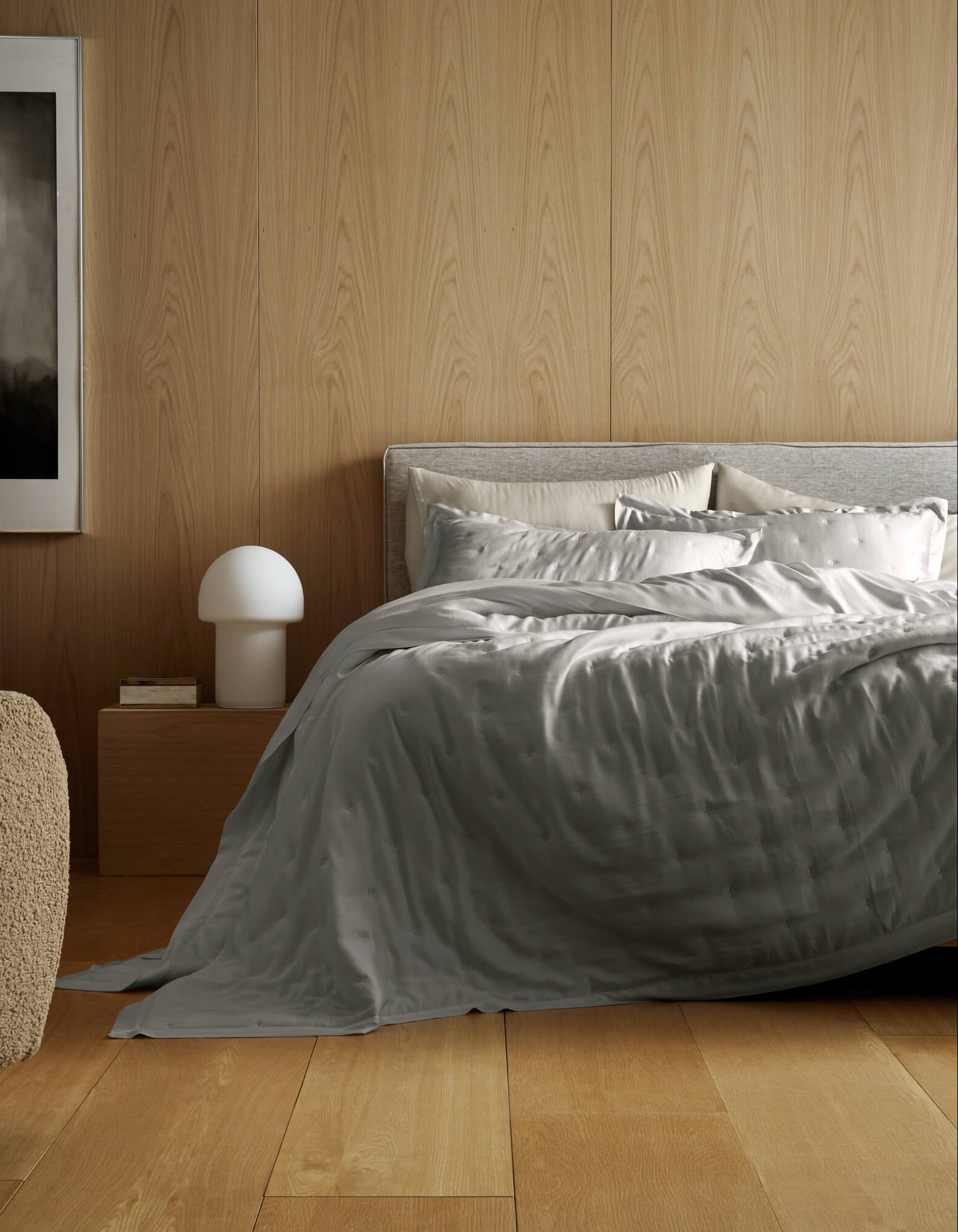 A modern bedroom featuring a light wood floor and wall, a neatly made bed with the Cozy Earth Bamboo Quilt Bundle in silver-gray, a wooden nightstand with a white lamp, a beige textured chair, and abstract wall art.