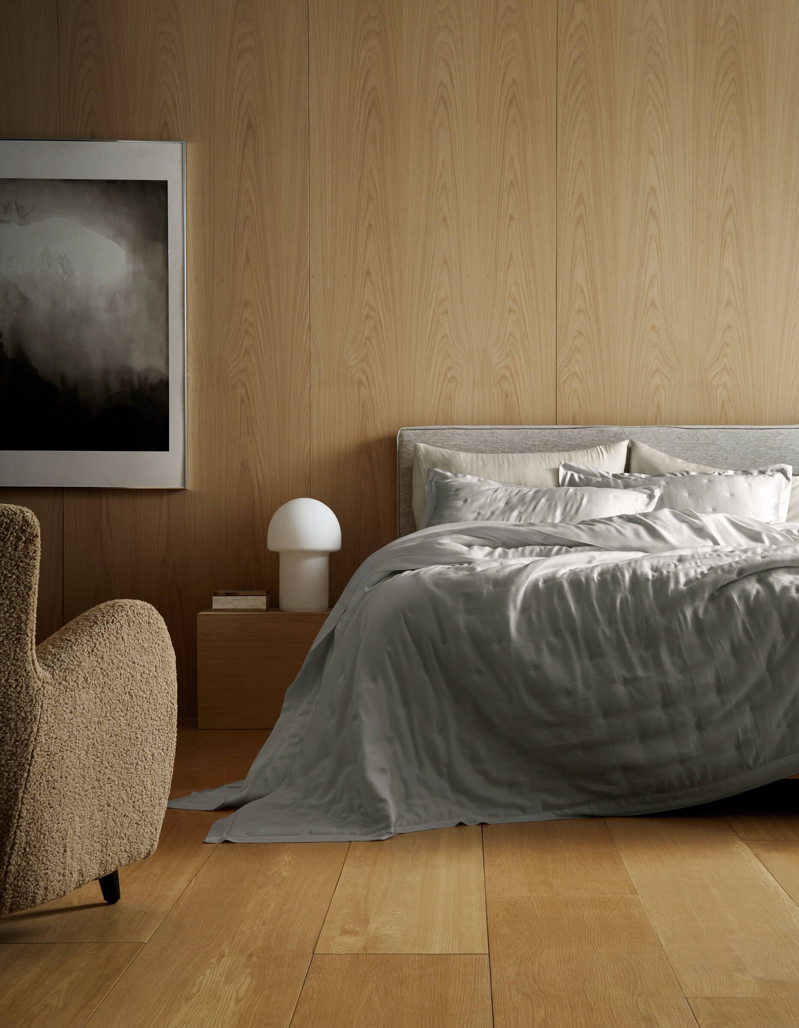 A modern bedroom with light wood walls and floors, a bed featuring Cozy Earth’s Bamboo Quilt in gray satin, a beige textured armchair, a mushroom-shaped white lamp on a wooden nightstand, and an abstract black-and-white painting.