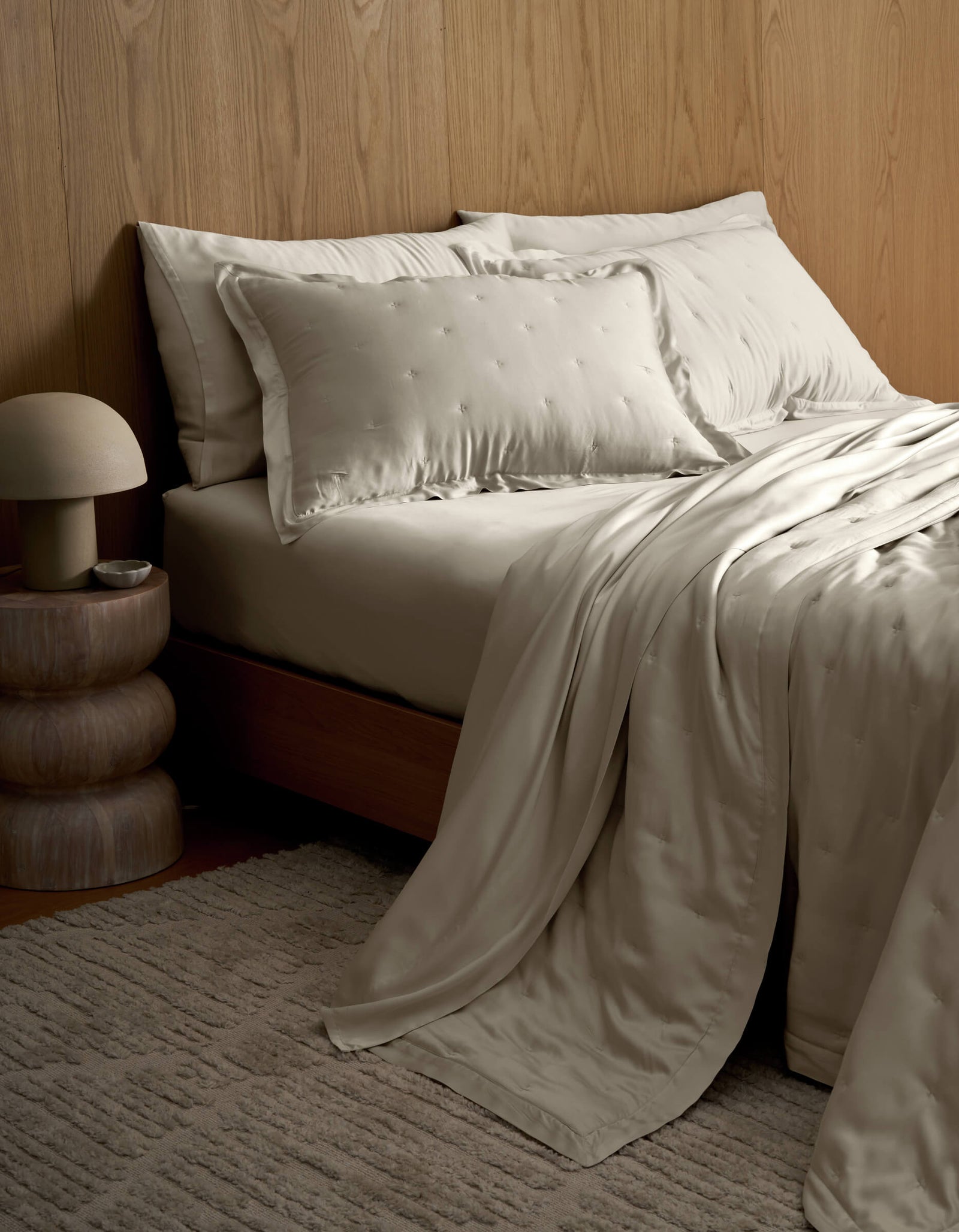 A neatly made bed featuring Cozy Earth's Bamboo Quilt with satin pillows sits beside a wooden nightstand with a round lamp, all set against a wood-paneled wall and a textured beige rug.