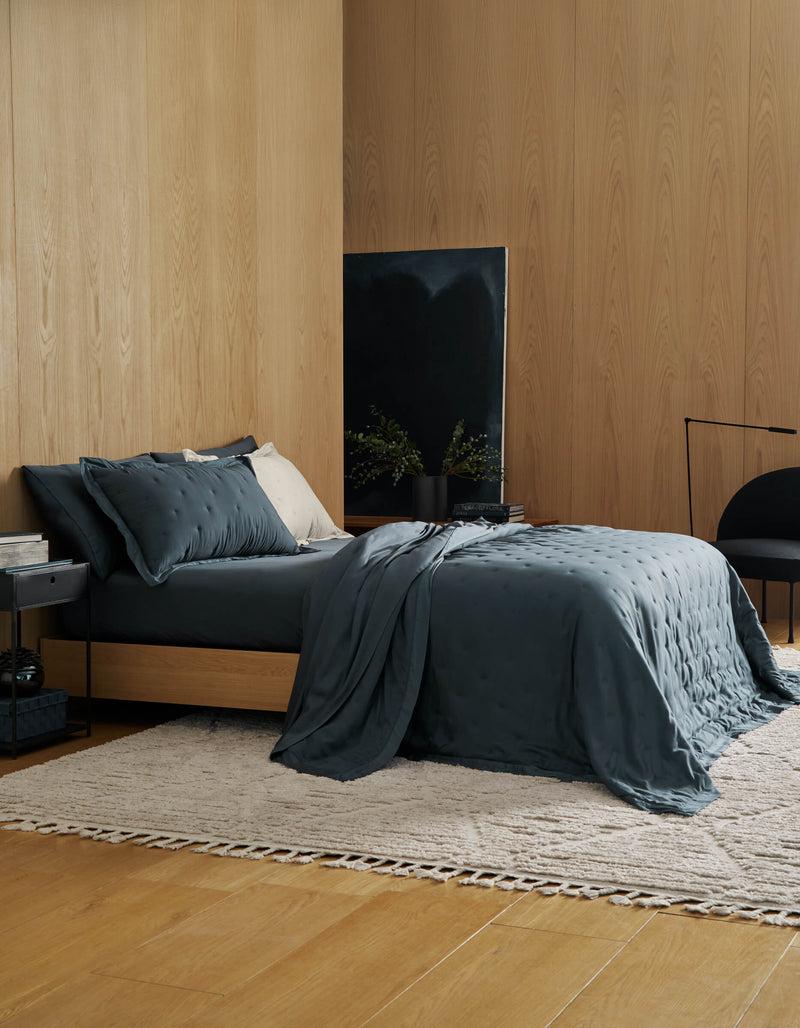 A modern bedroom with light wood walls and floor features a dark gray bed dressed in the Cozy Earth Bamboo Quilt, a small side table, light rug, black chair, and a dark abstract painting behind lush plants in a black vase.