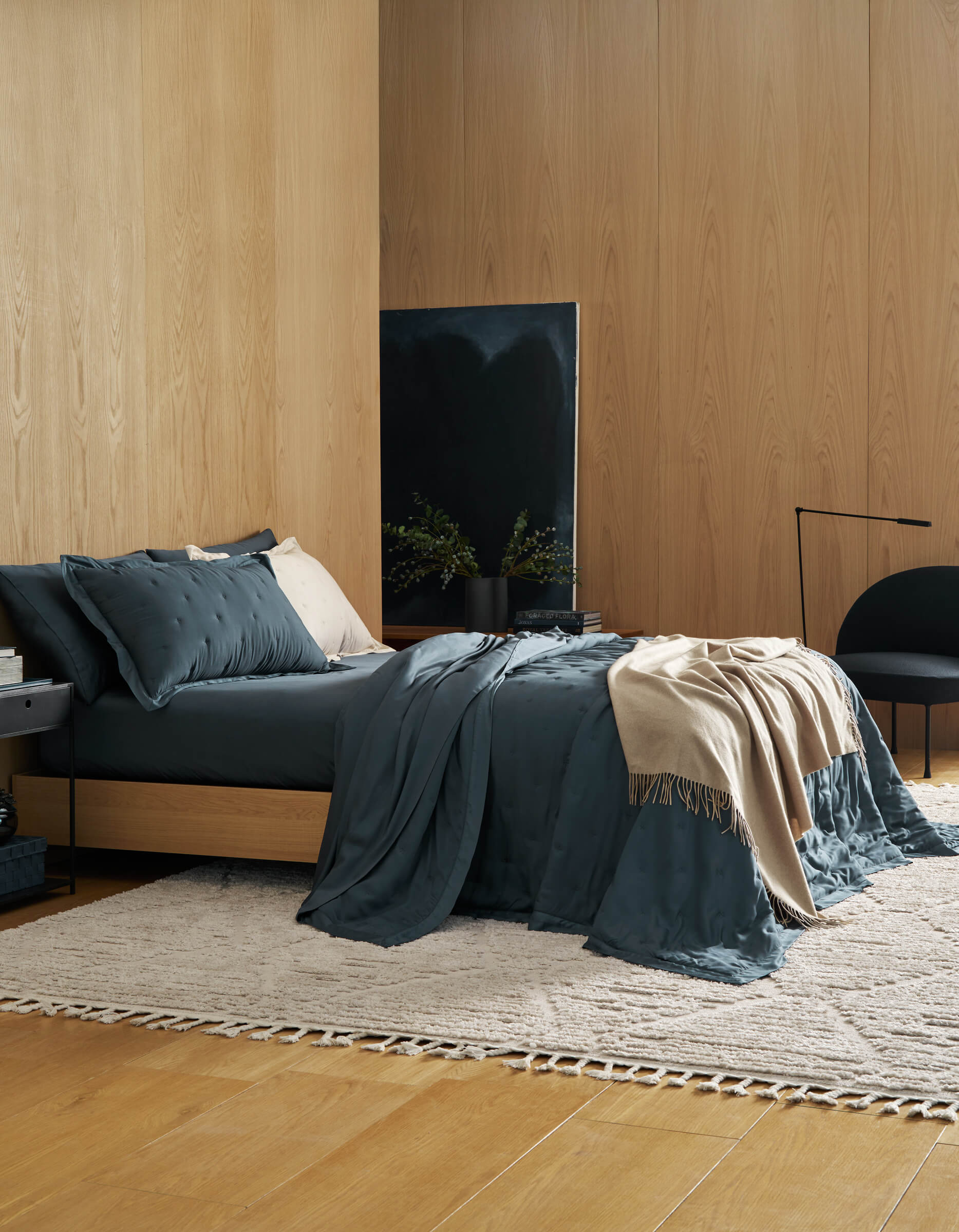 A modern bedroom features wood-paneled walls, a low bed with Cozy Earth's Bamboo Quilt Bundle in dark teal, a cream throw, and pillows. A neutral woven rug, black chair, side table, and minimalist decor create a cozy, stylish space.|Color:Pacific Blue
