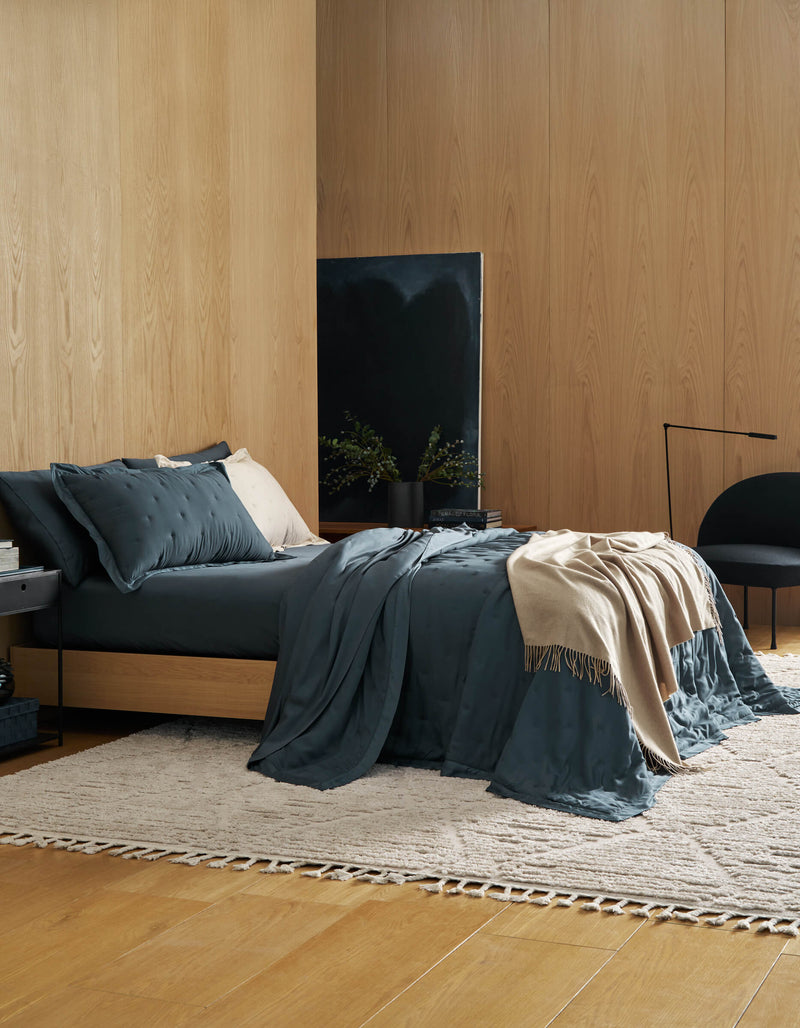 A modern bedroom features wood-paneled walls, a low bed with Cozy Earth's Bamboo Quilt Bundle in dark teal, a cream throw, and pillows. A neutral woven rug, black chair, side table, and minimalist decor create a cozy, stylish space.