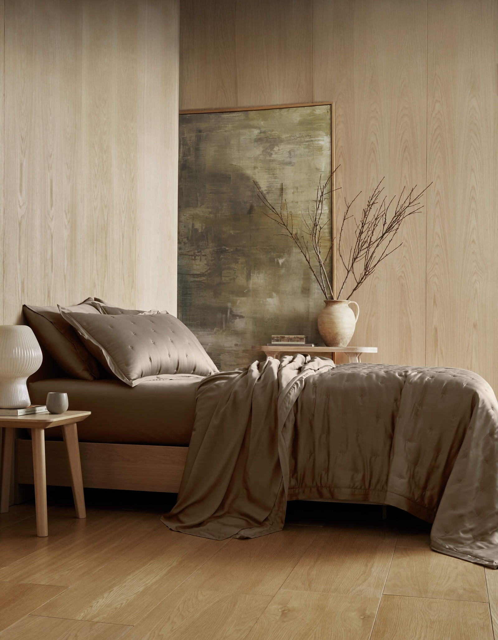 A minimalist bedroom with light wood walls and floor, a bed dressed in Cozy Earth’s Bamboo Quilt, a bedside table topped with a white lamp, and a large vase of bare branches set before an abstract painting.