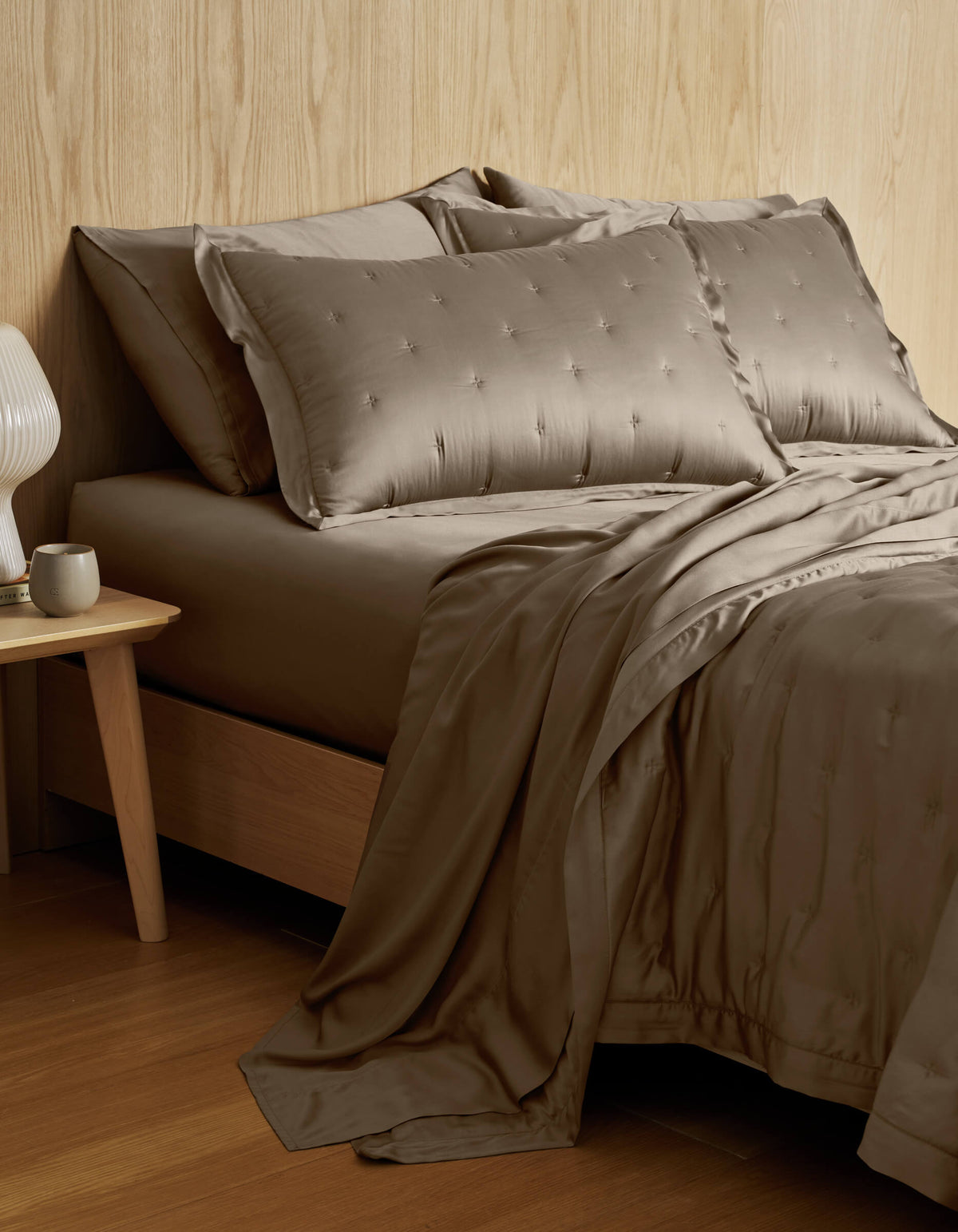 A neatly made bed featuring the Cozy Earth Bamboo Quilt and matching pillows in taupe sits beside a wooden nightstand with a white cup and modern lamp, set against a light wood-paneled wall.|Color:Walnut