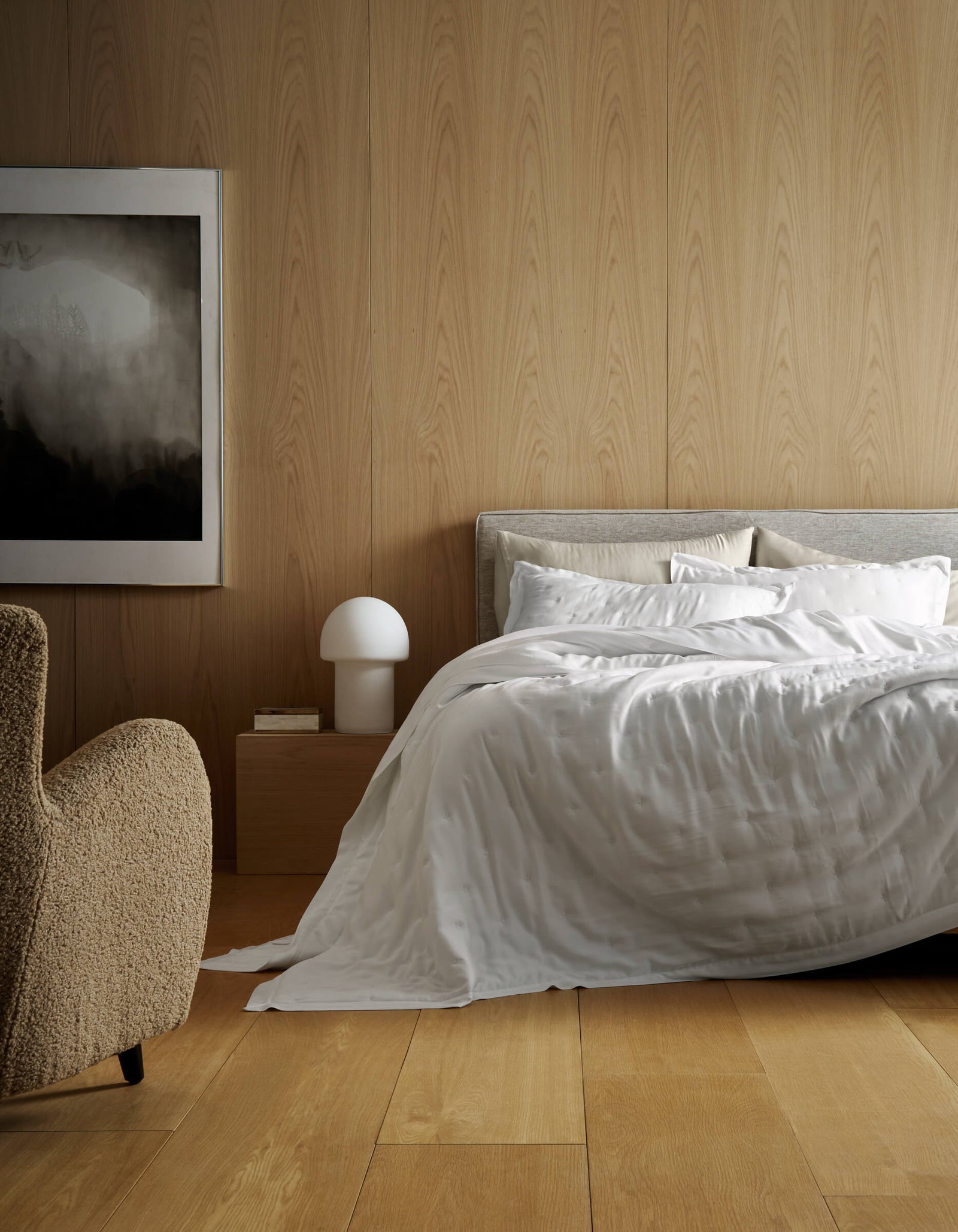 A modern bedroom features light wood panels, a bed made with Cozy Earth’s Bamboo Quilt, a beige armchair, a wooden nightstand with a white lamp, and abstract black-and-white artwork on the wall.