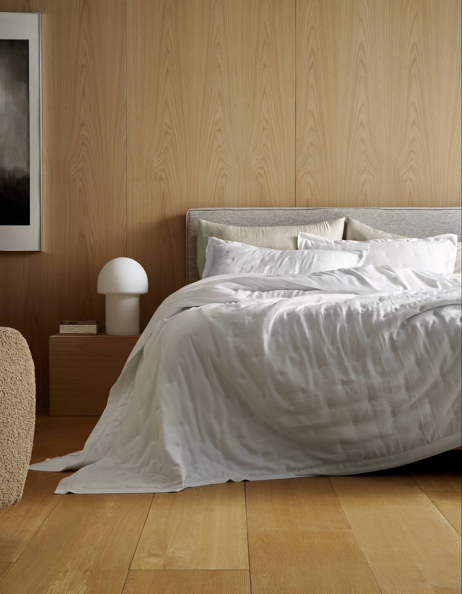 A modern bedroom features light wood-paneled walls, a large bed with Cozy Earth’s Bamboo Quilt Bundle, a beige chair, wooden nightstand with a white mushroom lamp, books, and an abstract painting on the wall.