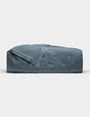The Cozy Earth Bamboo Quilt, shown folded against a plain white background, features a blue-gray color with a subtle quilted pattern and smooth, shiny finish.|Color:Pacific Blue