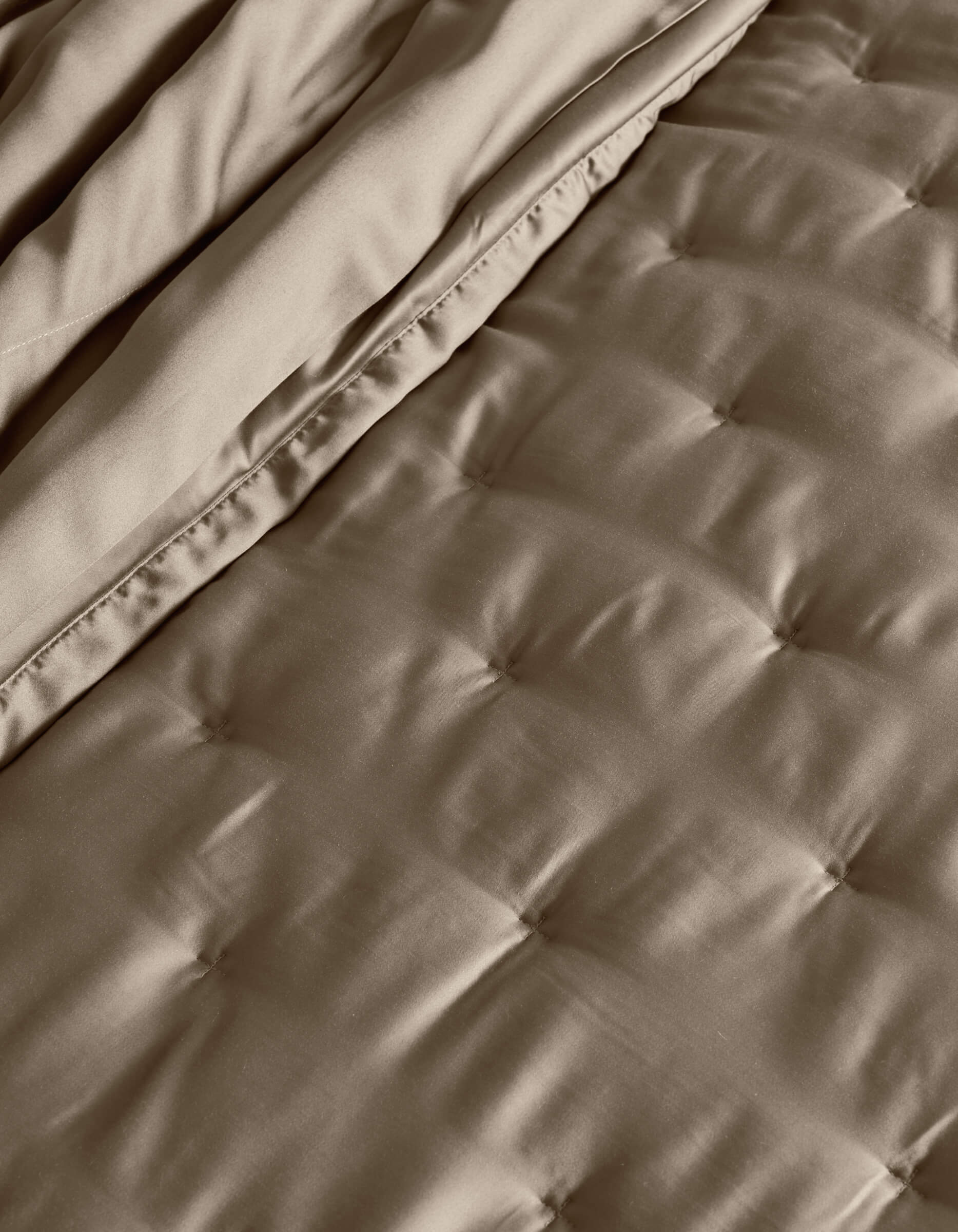 A close-up of the Cozy Earth Bamboo Quilt shows its quilted, taupe surface with stitched square patterns and a folded edge, highlighting the soft, smooth fabric texture.