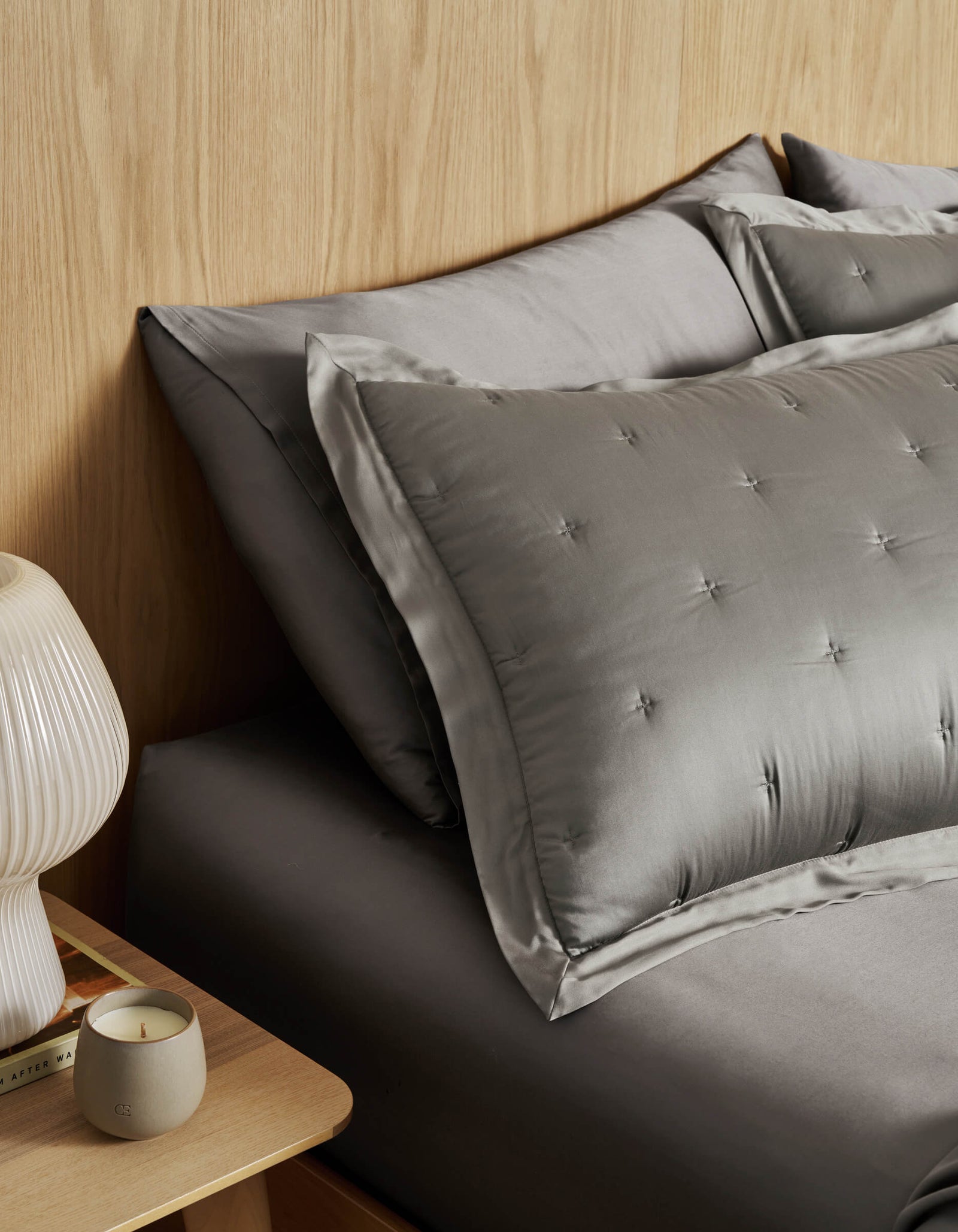 A neatly made bed with Cozy Earth's Bamboo Quilted Shams and tufted pillows sits beside a wooden nightstand with a ribbed white lamp, a candle, and books, set against a light wood-paneled wall.
