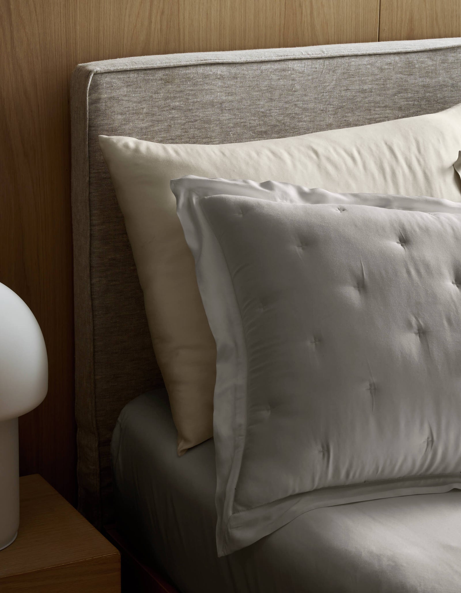 A neatly made bed with Cozy Earth Bamboo Quilted Shams, a padded fabric headboard, and a gray sheet. A modern white lamp is on a wooden nightstand beside the bed.