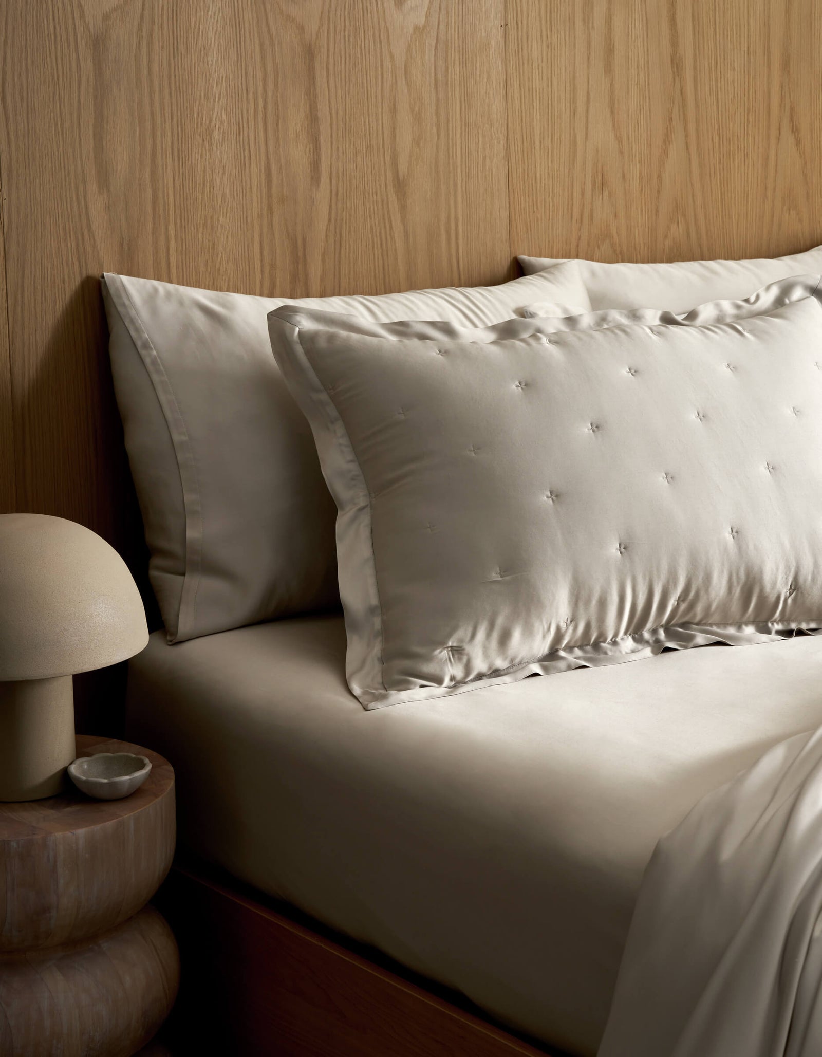 A neatly made bed features Cozy Earth's Bamboo Quilted Shams in light tones, set against a wooden headboard. A round bedside table holds a dome-shaped lamp and a small dish.
