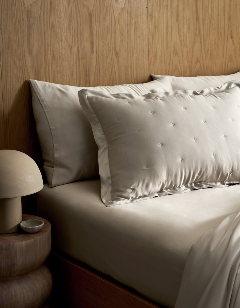 A neatly made bed features Cozy Earth's Bamboo Quilted Shams in light tones, set against a wooden headboard. A round bedside table holds a dome-shaped lamp and a small dish.