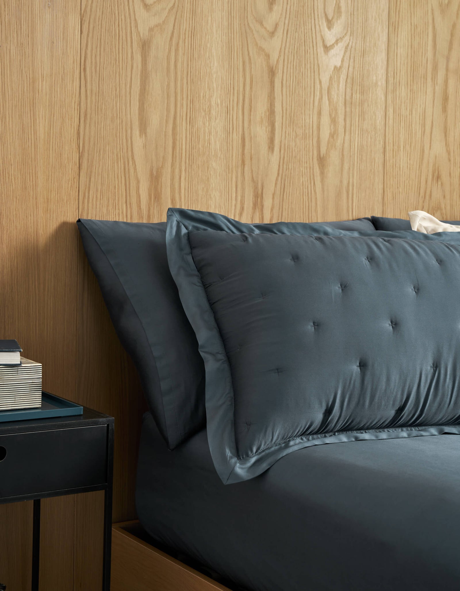 A neatly made bed with Cozy Earth Bamboo Quilted Shams in dark gray and matching sheets sits against a light wood-paneled wall. A black nightstand with stacked books is next to the bed.