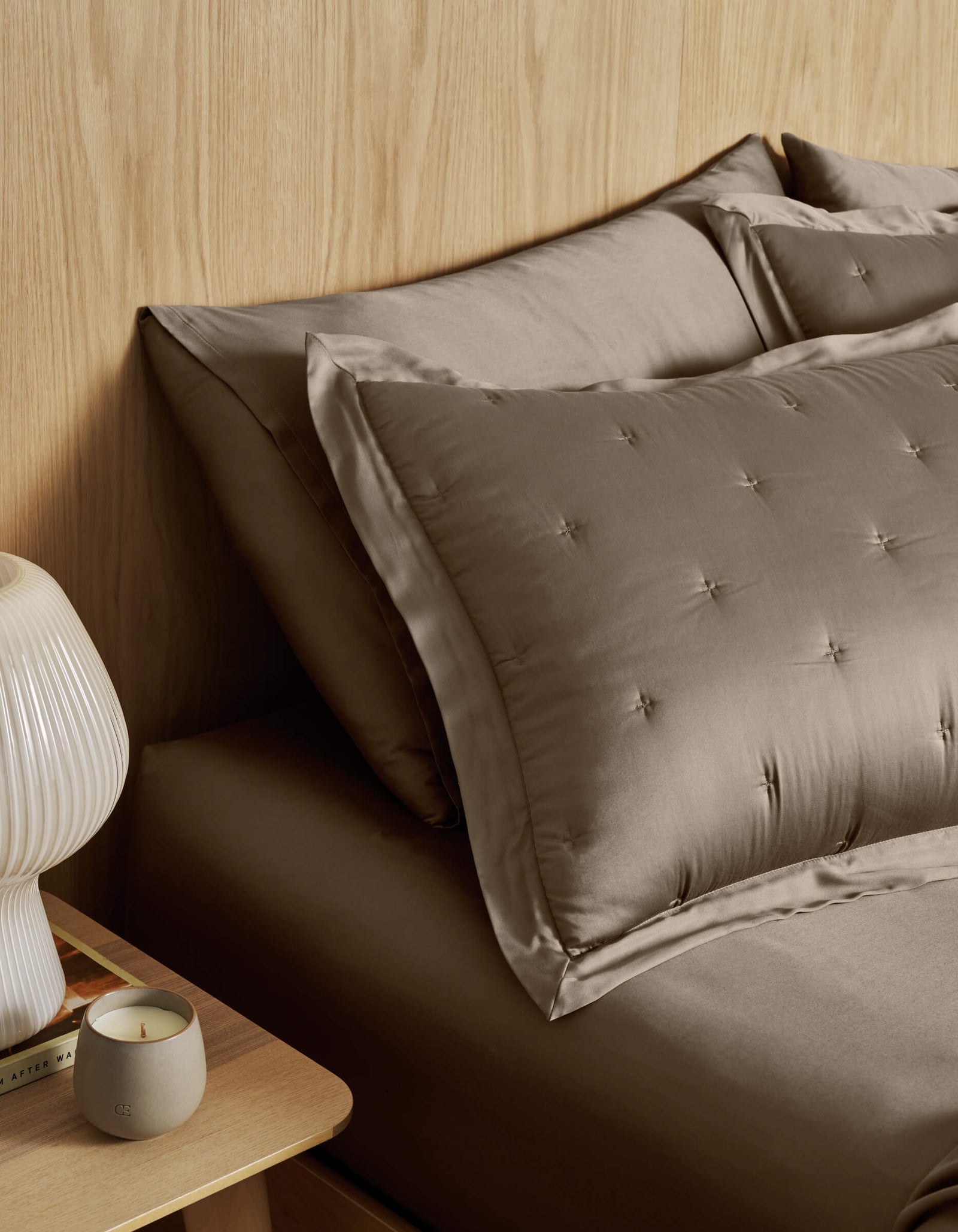 A neatly made bed with Cozy Earth Bamboo Quilted Shams and taupe satin bedding sits by a wooden nightstand topped with a ribbed white lamp, a lit candle, and a book against a light wood-paneled wall.