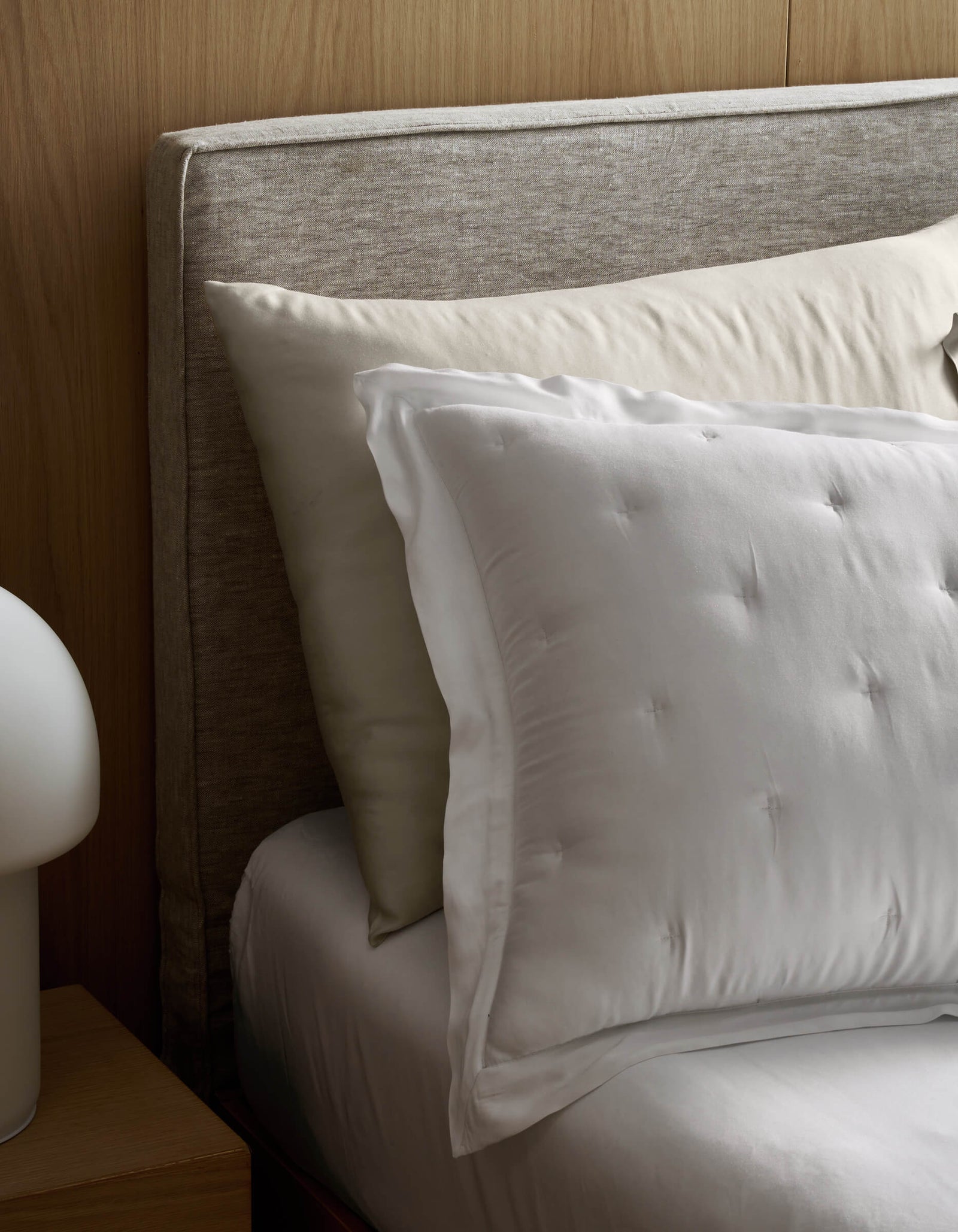 A close-up of a bed featuring Cozy Earth Bamboo Quilted Shams, paired with cream and white pillows, white sheets, a light gray upholstered headboard, and part of a round white bedside lamp on a wooden nightstand.