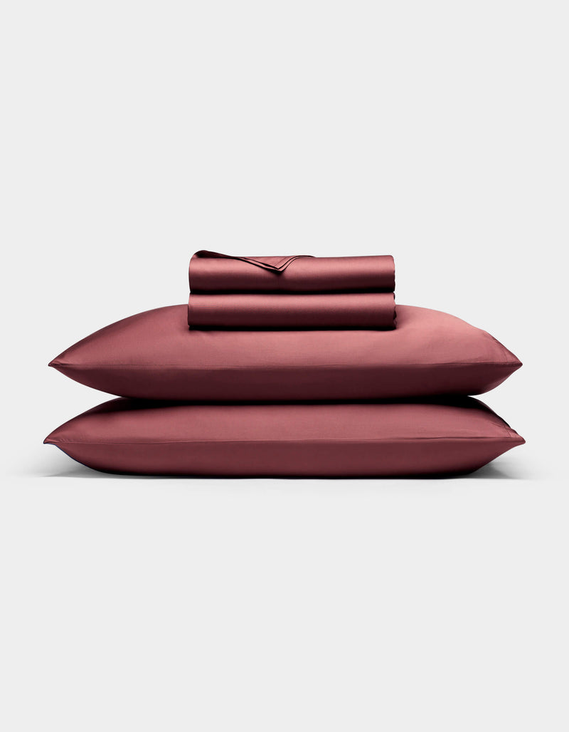 A neatly folded Bamboo Sheet Set by Cozy Earth, including bed sheets and pillowcases in deep burgundy, is stacked on two matching pillows against a plain light gray background.