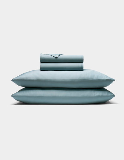 The Cozy Earth Bamboo Sheet Set, featuring two pillows and a folded sheet in pale blue, is neatly stacked against a plain white background.|Color:River