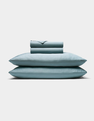 The Cozy Earth Bamboo Sheet Set, featuring two pillows and a folded sheet in pale blue, is neatly stacked against a plain white background.|Color:River