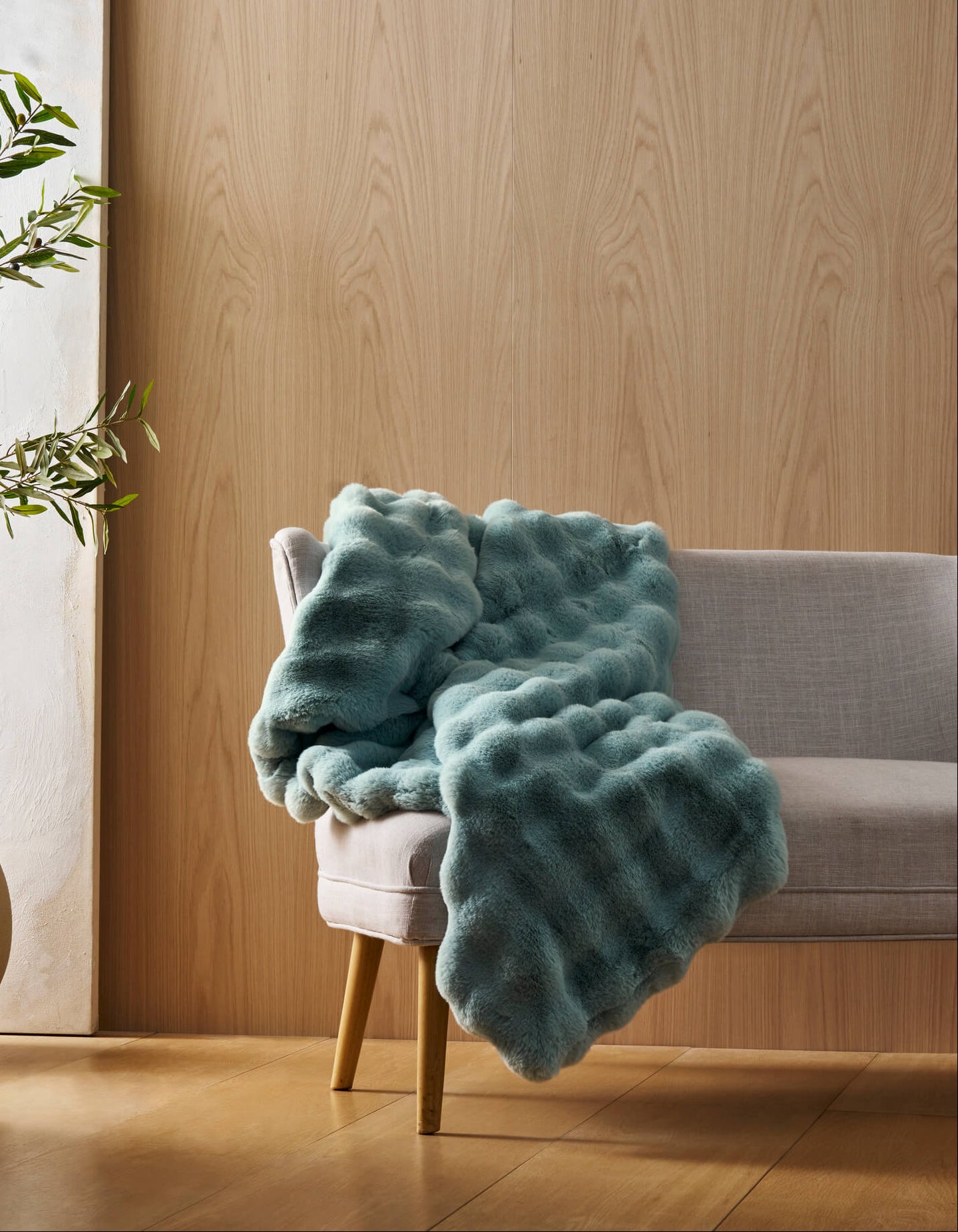 A light gray chair with wooden legs displays the Bubble Cuddle Blanket by Cozy Earth in textured teal, set in a room with light wood-paneled walls, a potted plant, and a white canvas. Natural light brightens the space.