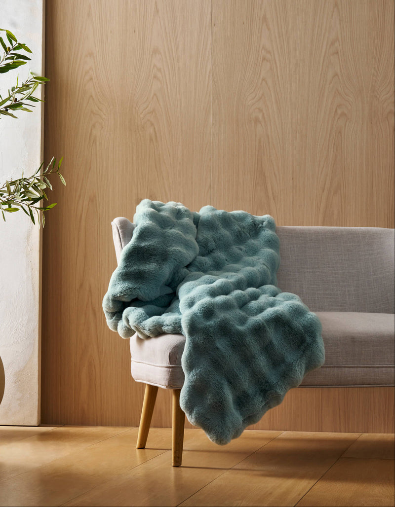 A light gray chair with wooden legs displays the Bubble Cuddle Blanket by Cozy Earth in textured teal, set in a room with light wood-paneled walls, a potted plant, and a white canvas. Natural light brightens the space.
