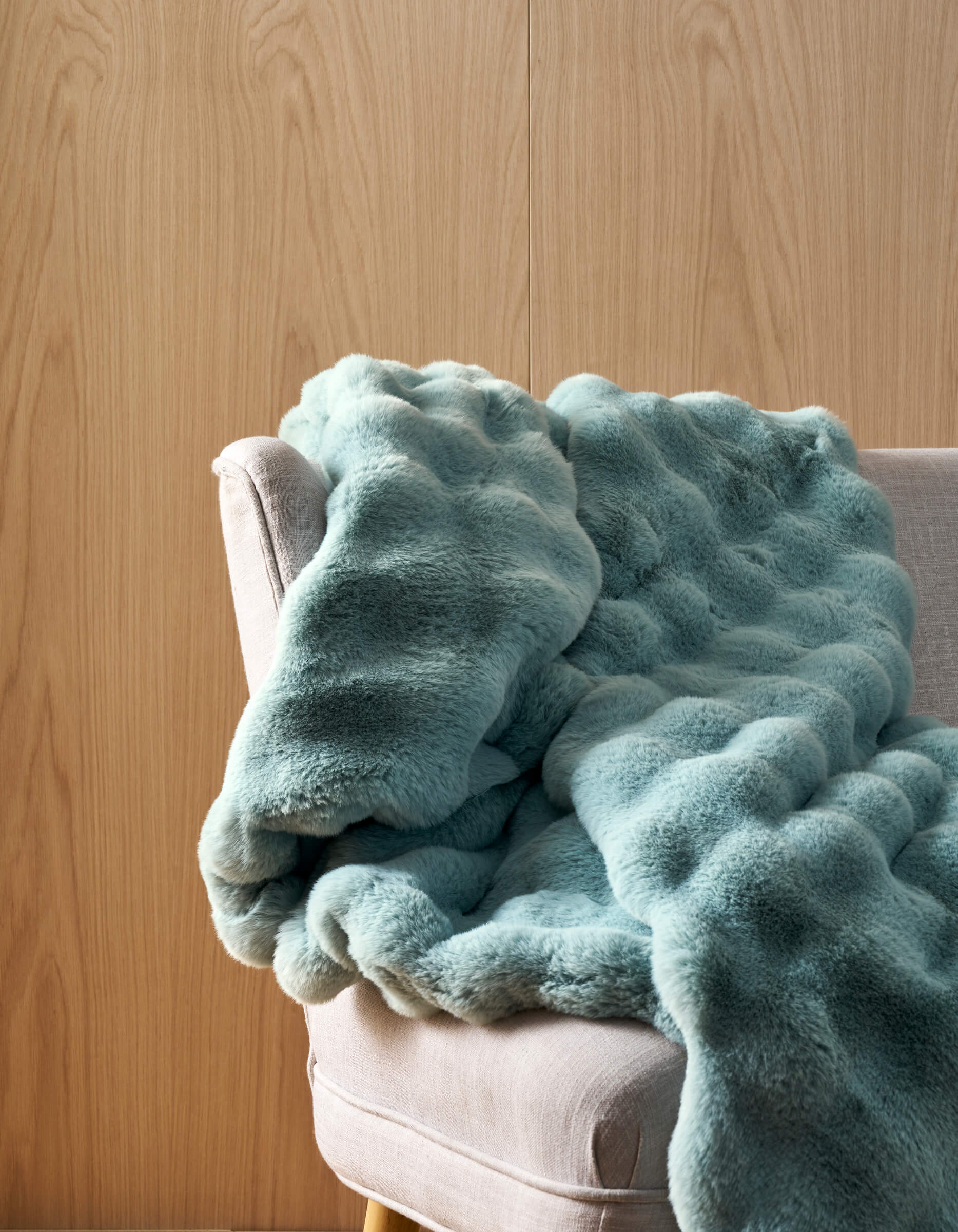The Cozy Earth Bubble Cuddle Blanket, featuring plush light blue faux fur with a wavy texture, is draped over a light gray upholstered chair set against a light wood-paneled wall.|Color:Tide