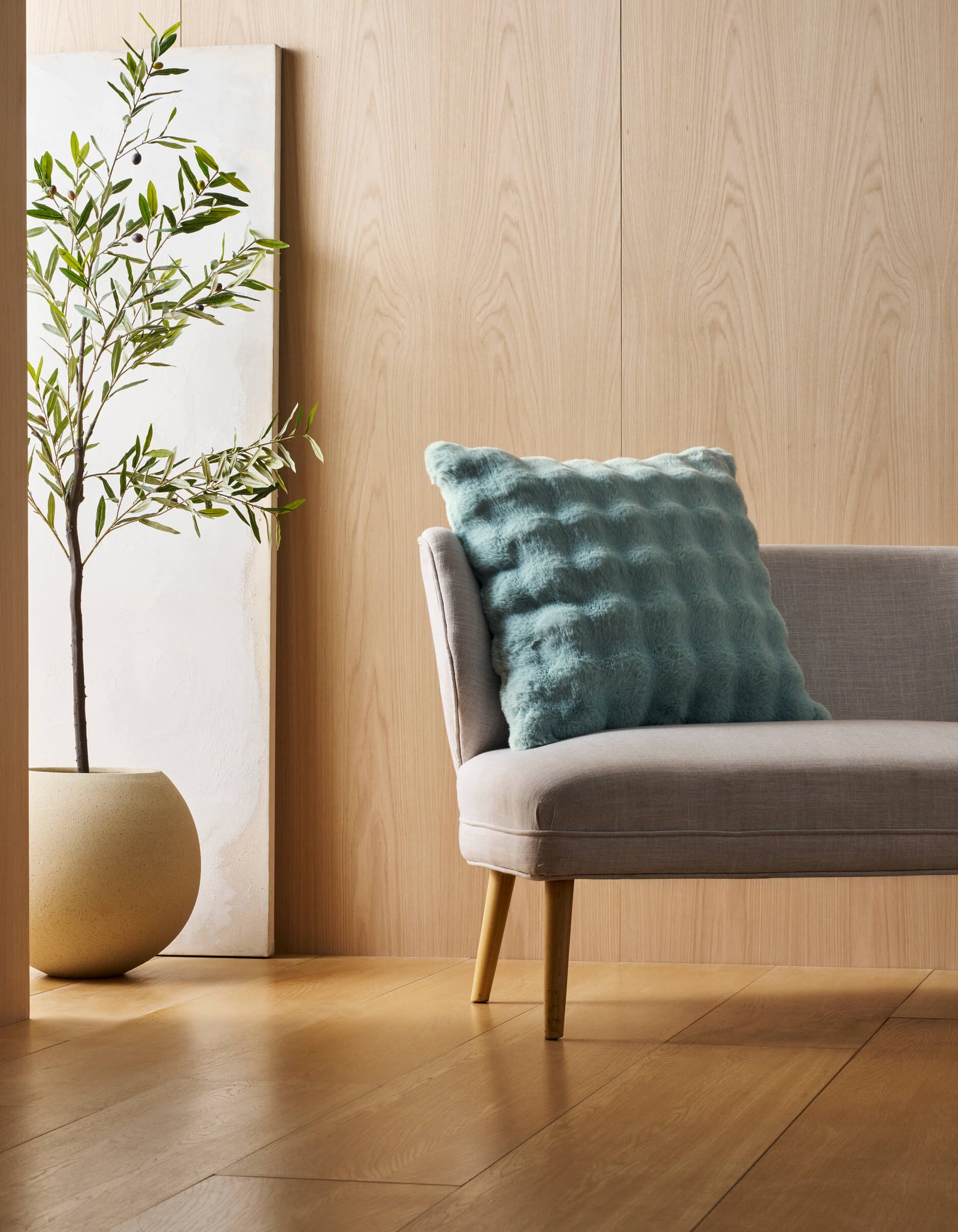 A modern, light gray chair with wooden legs sits on a wooden floor. A Cozy Earth Bubble Cuddle Pillow rests on the chair. Nearby, an olive tree in a round pot and a tall canvas lean against a light wood-paneled wall.
