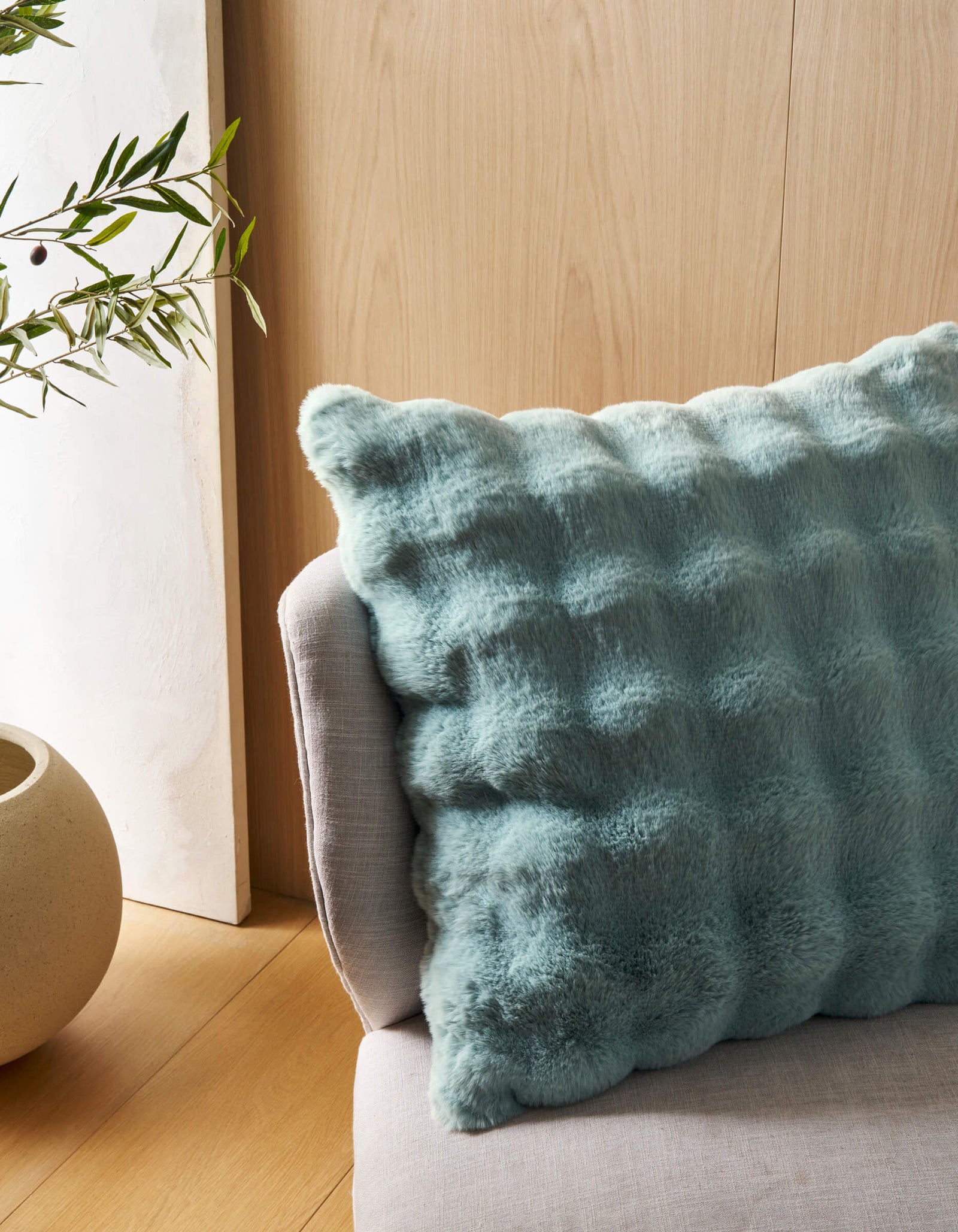 A close-up of the Cozy Earth Bubble Cuddle Pillow in light blue, with a fluffy texture, rests on a light grey sofa beside a round beige planter with green leafy branches, set against a light wood-paneled wall.