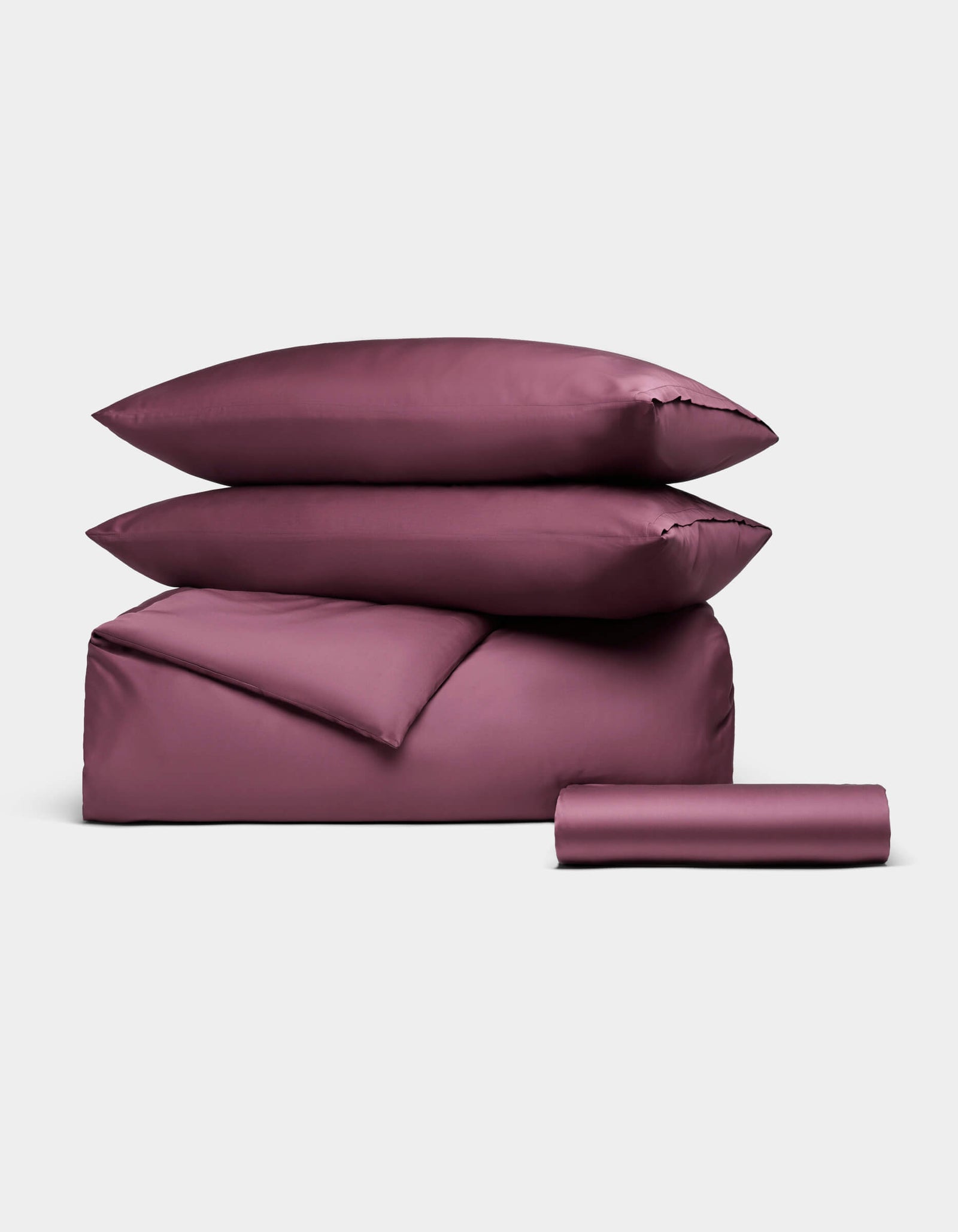 The Cozy Earth Bamboo Bedding Essential Bundle is shown neatly folded, featuring two stacked pillows, a folded duvet, and a rolled sheet in mauve, all set against a plain light gray background.