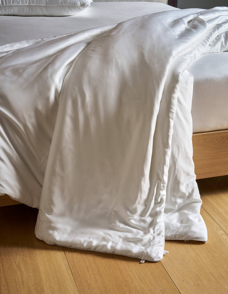 A Cozy Earth Silk Comforter drapes over the edge of a light wooden bed frame onto a wooden floor, with matching white bedding visible on the bed.