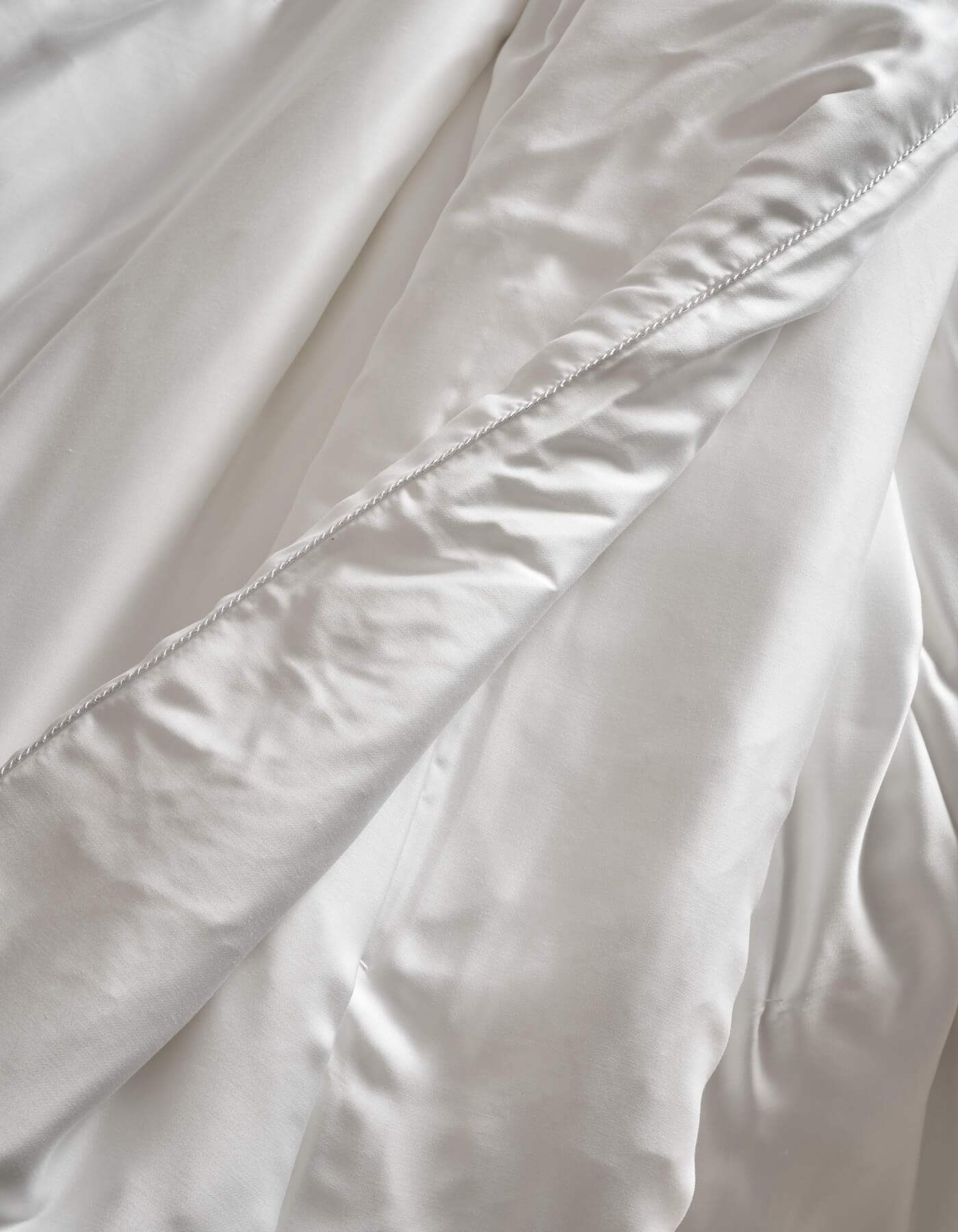 Close-up of Cozy Earth Silk Comforter: soft, white fabric with a diagonal seam, gentle folds, and smooth, shiny texture.