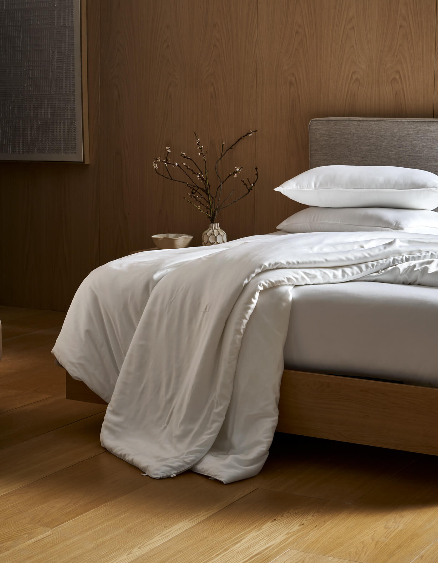 A neatly made bed with a gray headboard and white bedding features the Cozy Earth Silk Comforter draped over the edge. A vase with branches sits on the bedside table, all set on wooden floors with a wood-paneled wall in the background.