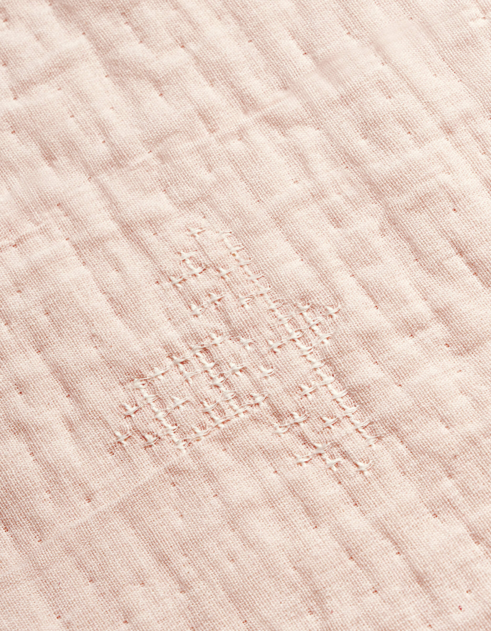 A close-up of the Cozy Earth Little Bee Matelasse Baby Blanket shows its white fabric with a grid-like, hand-stitched repair in matching thread, highlighting the textured and slightly uneven surface.|Color:Petal