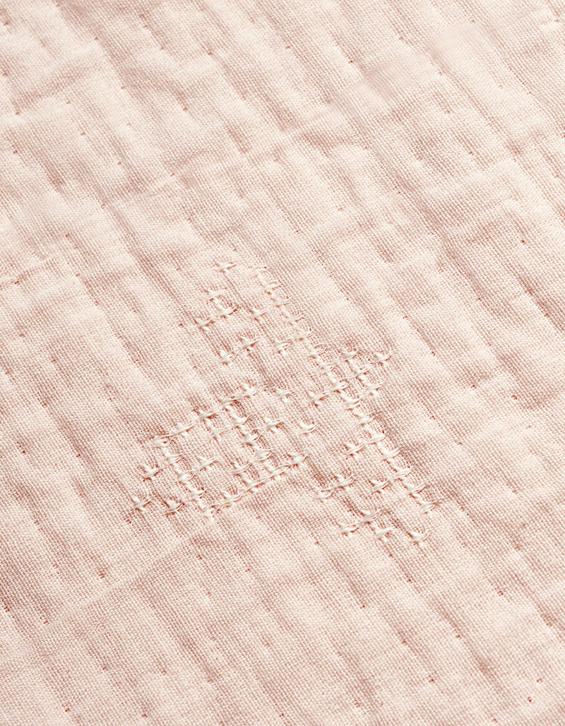 A close-up of the Cozy Earth Little Bee Matelasse Baby Blanket shows its white fabric with a grid-like, hand-stitched repair in matching thread, highlighting the textured and slightly uneven surface.