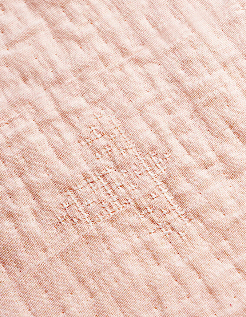 A close-up of the Cozy Earth Little Bee Matelasse Baby Blanket shows its white fabric with a grid-like, hand-stitched repair in matching thread, highlighting the textured and slightly uneven surface.