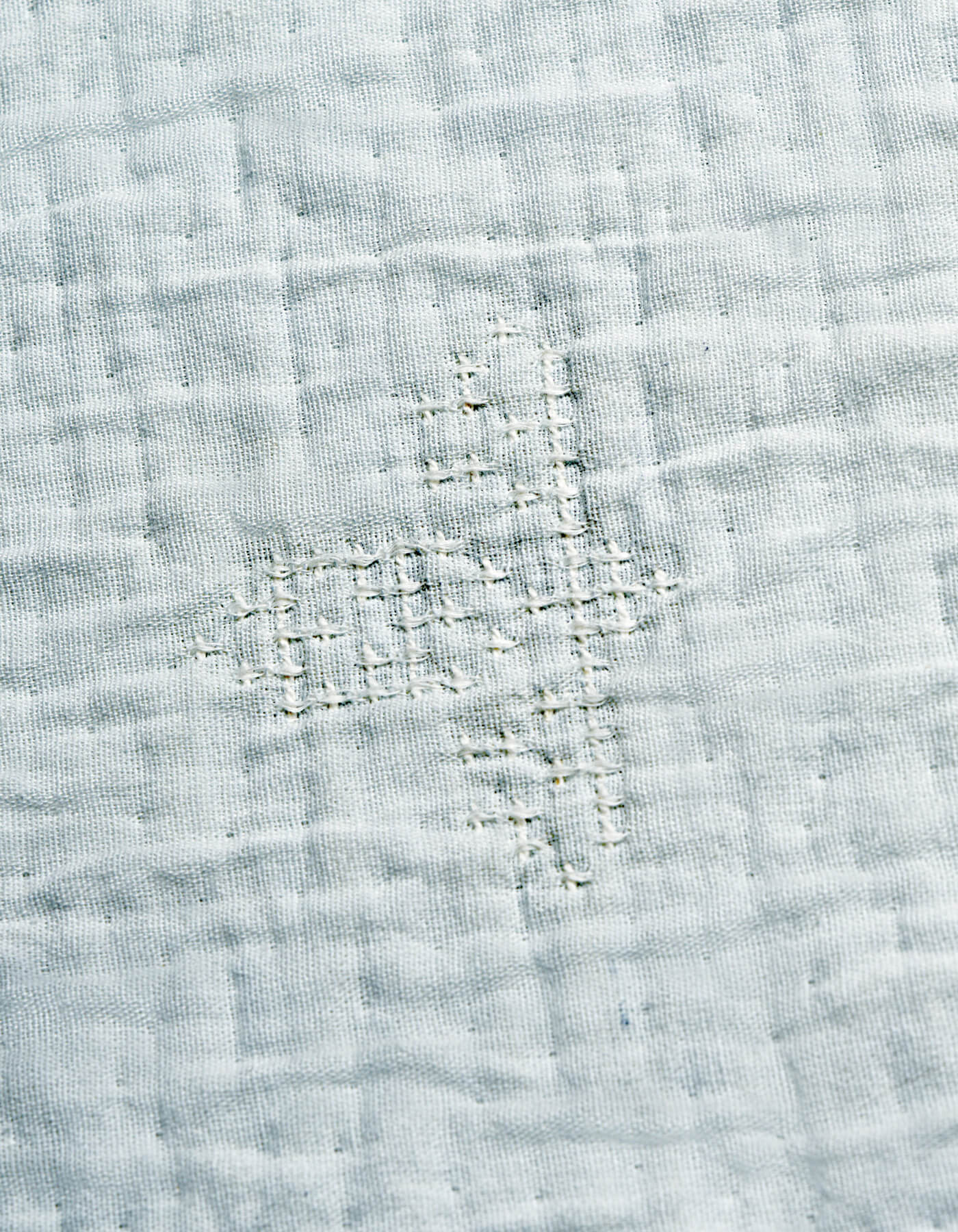 A close-up of the Cozy Earth Little Bee Matelasse Baby Blanket shows its white fabric with a grid-like, hand-stitched repair in matching thread, highlighting the textured and slightly uneven surface.