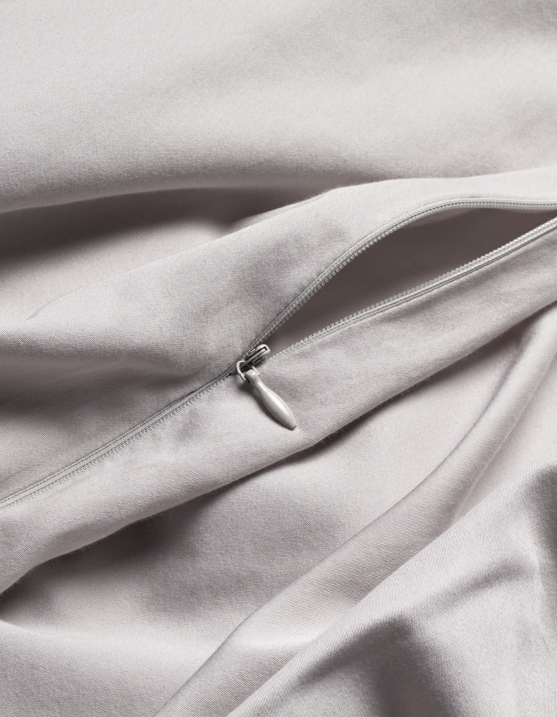 A close-up of Cozy Earth's Soft-Wash Cotton Duvet Cover in light grey shows a silver zipper partially unzipped on its smooth, gently folded fabric.